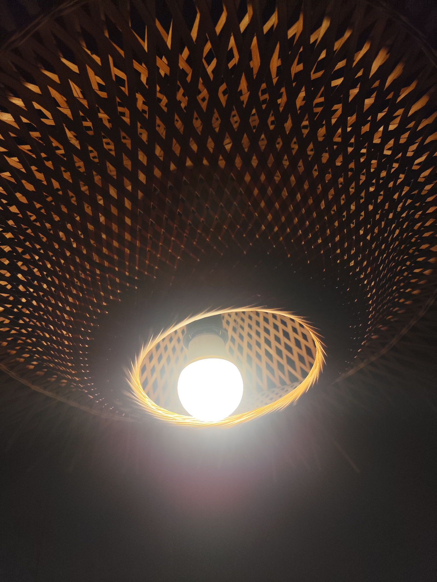 A low-angle shot reveals a lightbulb shining brightly inside a woven, basket-like lampshade in Da Lat, Vietnam. The shade is constructed from light brown, woven strips, forming a diamond-like pattern. The lightbulb is positioned at the center of the shade. The background is dark and out of focus.