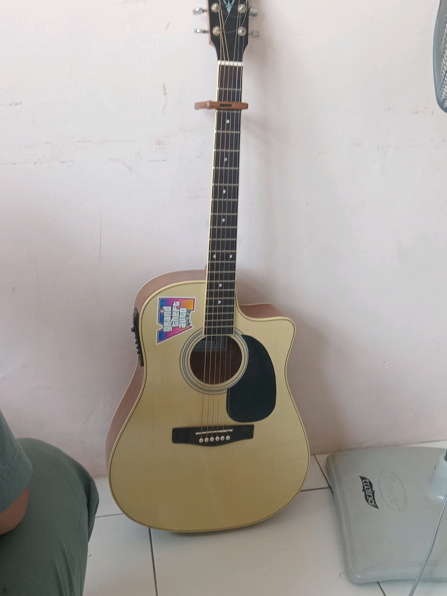 An acoustic guitar is leaning against a white wall. The guitar is light tan with a black pickguard and silver tuning pegs. It has a wooden capo attached to the neck and a colorful sticker on the body. To the left of the guitar, a person's legs are visible, wearing green pants. On the right side of the image, a portion of a white electric fan is visible on the floor. The floor is tiled. The scene appears to be indoors, possibly in a residence. The image does not contain enough information to determine its location in Kalikajar, Indonesia.