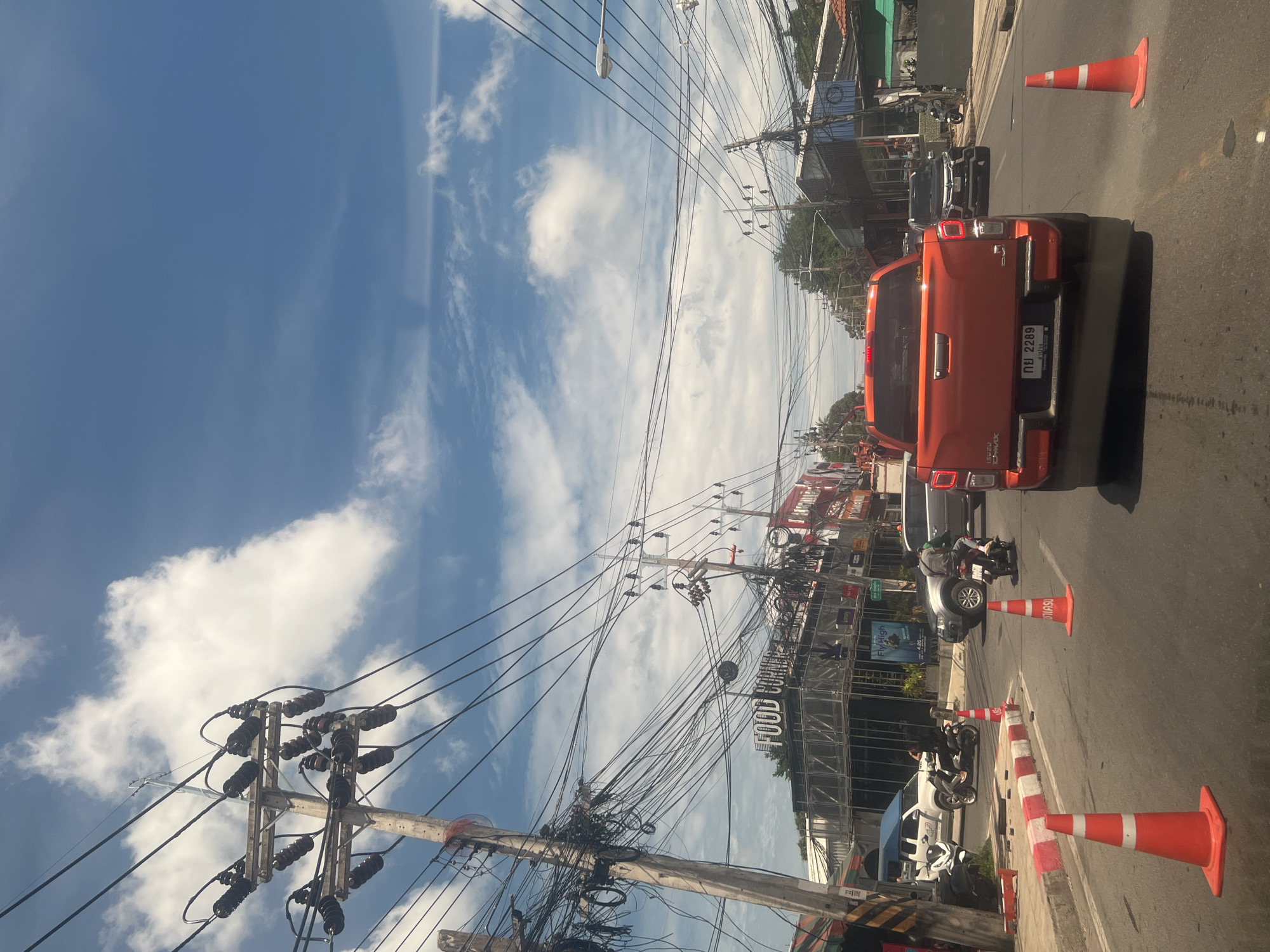 In the city of Ko Samui District, Thailand, this street scene unfolds under a bright, partly cloudy sky. The road is filled with vehicles, including an orange pickup truck in the foreground with a license plate reading "กย 2289". To the left, a white SUV is parked, and a motorcycle passes by. Orange traffic cones are strategically placed on the road. Overhead, a dense network of electrical wires hangs precariously from utility poles, a common sight in many parts of Thailand. Buildings line the street, with one featuring a sign that reads "FOOD CORNER". Other vehicles, including cars and motorcycles, are visible in the background, suggesting moderate traffic flow. The overall impression is that of a bustling urban environment with the characteristic visual elements of Southeast Asian street life.