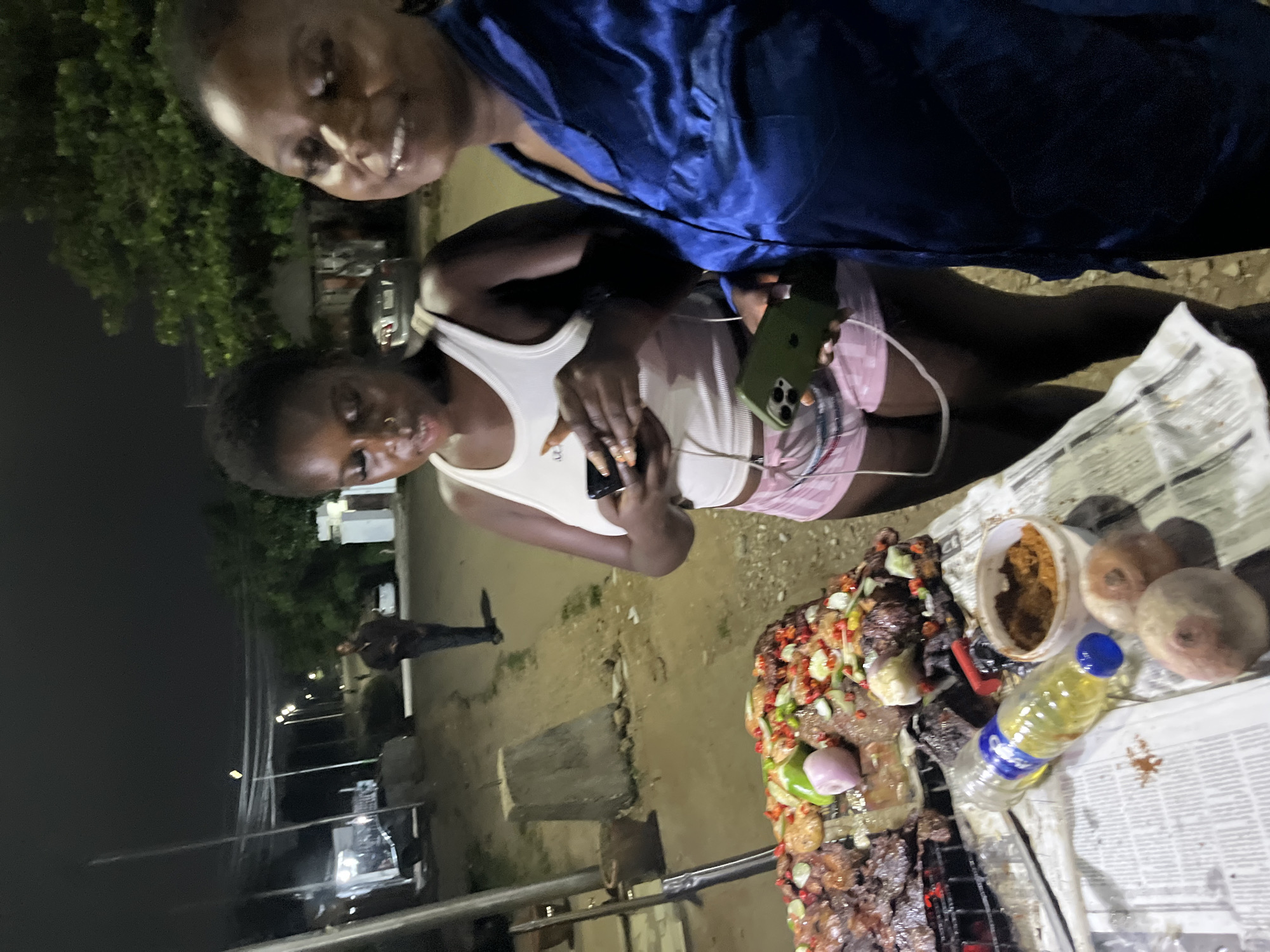 Two young women are standing next to a grill laden with barbecued meat and vegetables, in an outdoor setting at night, possibly in Abuja, Nigeria. The woman on the left is wearing a white tank top and pink plaid shorts, and is holding a smartphone. The woman on the right is wearing a blue satin robe and is smiling. The grill appears to be set up on a table covered with newspaper. In the background, there is a dirt road, streetlights, trees, and a few buildings. A man is walking in the distance. The scene suggests a casual outdoor meal or street food vendor.