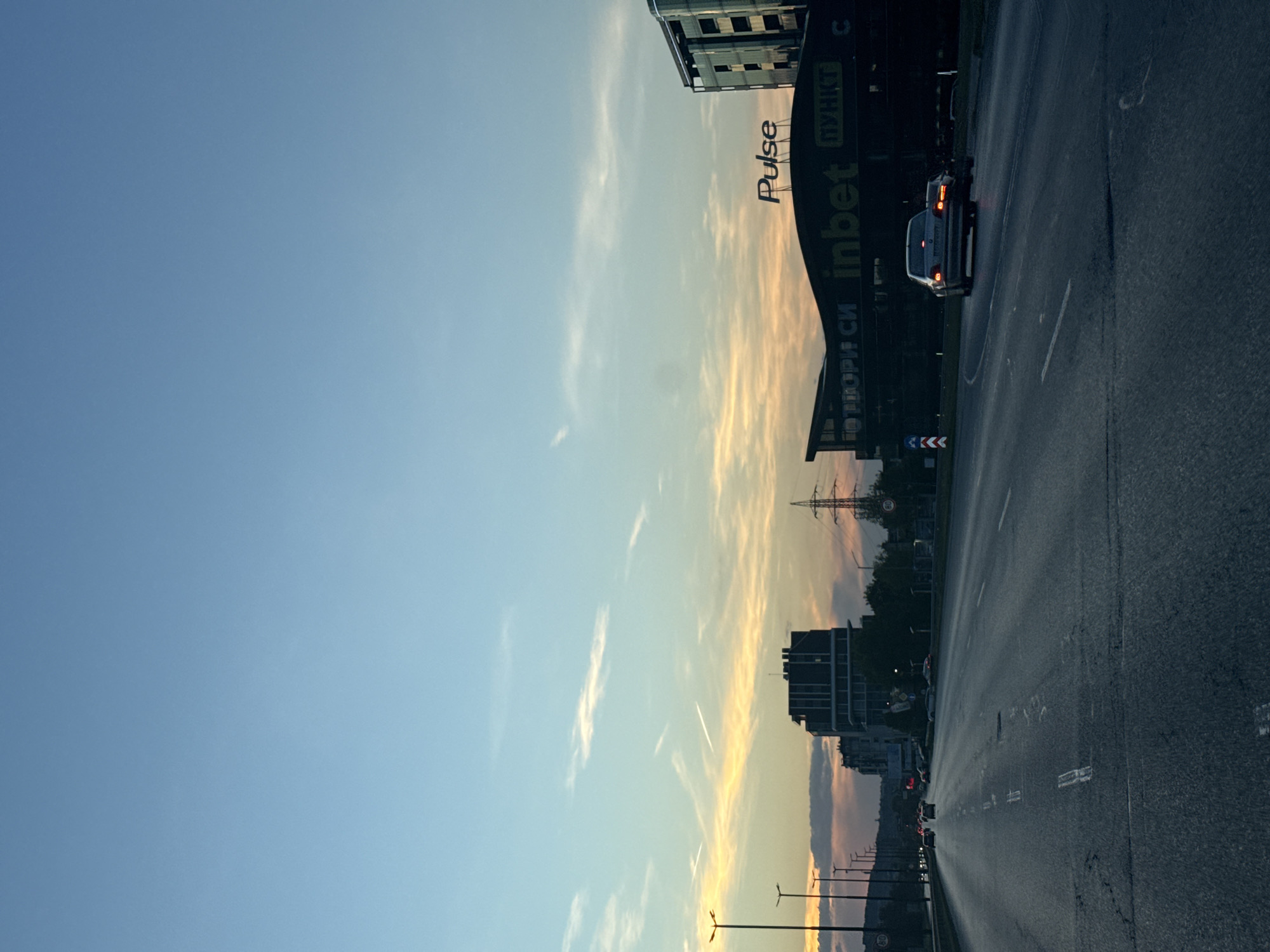 The image shows an urban landscape in Sofia, Bulgaria, at dusk or dawn, likely captured from a moving vehicle. A wide road stretches into the distance, with a car visible on the right side. Buildings are on either side of the road, including a large structure with "Pulse" and "inbet" signage. Streetlights line the road, and power lines are visible in the background. The sky is a blend of blue and orange, suggesting either sunrise or sunset.