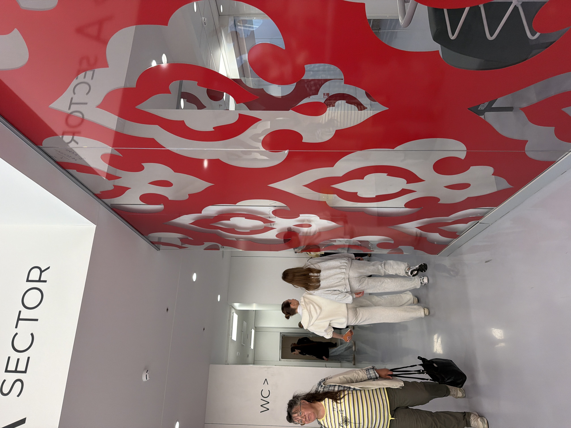In an indoor, brightly lit hallway with a polished gray floor, the words "SECTOR A" are visible in large black lettering on a white overhead sign. To the left, a woman with gray hair and glasses, wearing a yellow and white striped shirt and a beige jacket, walks towards the viewer, carrying a black handbag. Further down the hallway, three young women are walking away from the camera, two in light-colored sweat suits and one in a dark jacket. A sign reading "WC >" is mounted on the wall to the left. A large glass partition with a red and white ornamental pattern runs along the right side of the hallway.