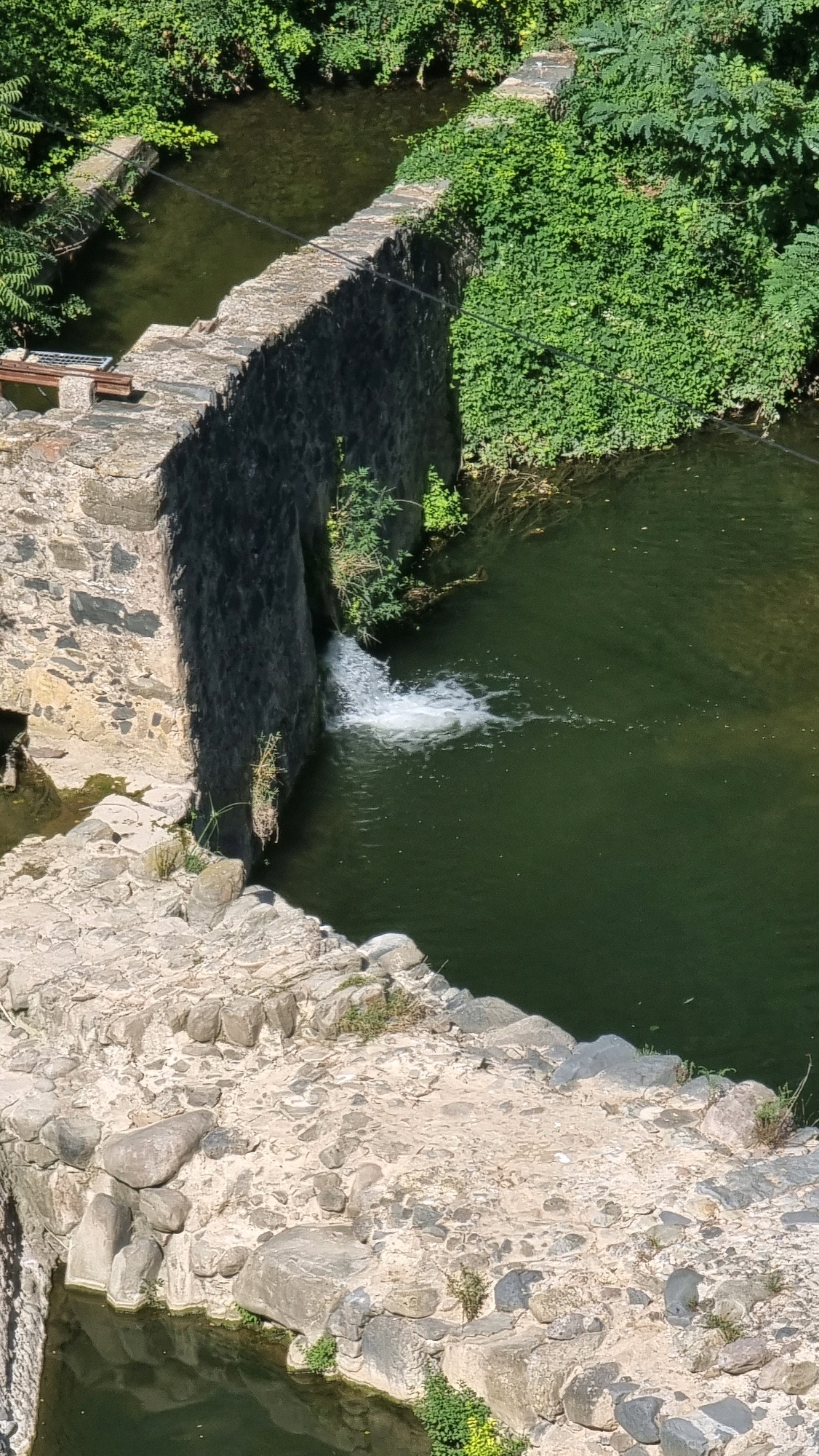 This is a medium-shot, high-angle, vertical outdoor photograph of a small, man-made waterfall in Castellfollit De La Roca, Spain. The waterfall flows from a stone wall structure into a dark green body of water. The surrounding environment is lush with green foliage. The stone walls are constructed with irregular, rough-hewn rocks. The water is murky and shows ripples from the falling water. There are no people or animals visible in the image. The scene appears to be a natural or semi-natural water feature integrated into an ancient stone structure, possibly an old mill or aqueduct. The lighting suggests a sunny day.