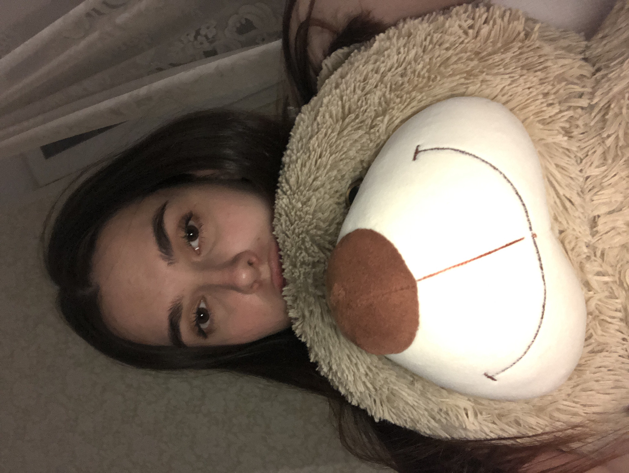 A young woman with dark hair is lying down, her face close to the camera. She is holding a large, tan-colored stuffed animal, possibly a bear, which is partially obscuring her view. The bear has a white face with a brown nose and stitched facial features. The background is blurry but appears to be a curtain or wall with a subtle pattern. The image is rotated, placing the woman and bear in a vertical orientation. There is no evidence in the image to confirm the location in the city of Noviy, Russia.