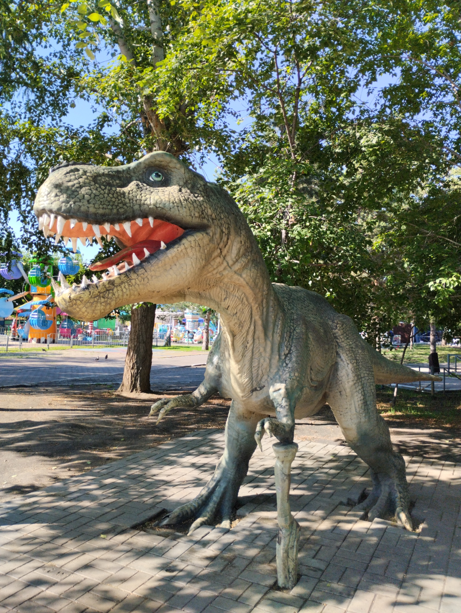 The image depicts a life-sized dinosaur model, likely a Tyrannosaurus Rex, standing on a paved walkway in an outdoor park or amusement area. The dinosaur is green and gray, with its mouth open revealing sharp teeth and a red tongue. It appears to be a static exhibit. In the background, there are trees with green foliage, a blue sky, and amusement park rides, including a Ferris wheel and colorful attractions. The ground is covered with light-colored paving stones, and shadows from the trees are cast across the walkway. The location is identified as Kostanay, Kazakhstan. There are no people visible in the immediate vicinity of the dinosaur, though some birds are on the ground in the mid-ground. The scene suggests a sunny day, with bright lighting and clear visibility.