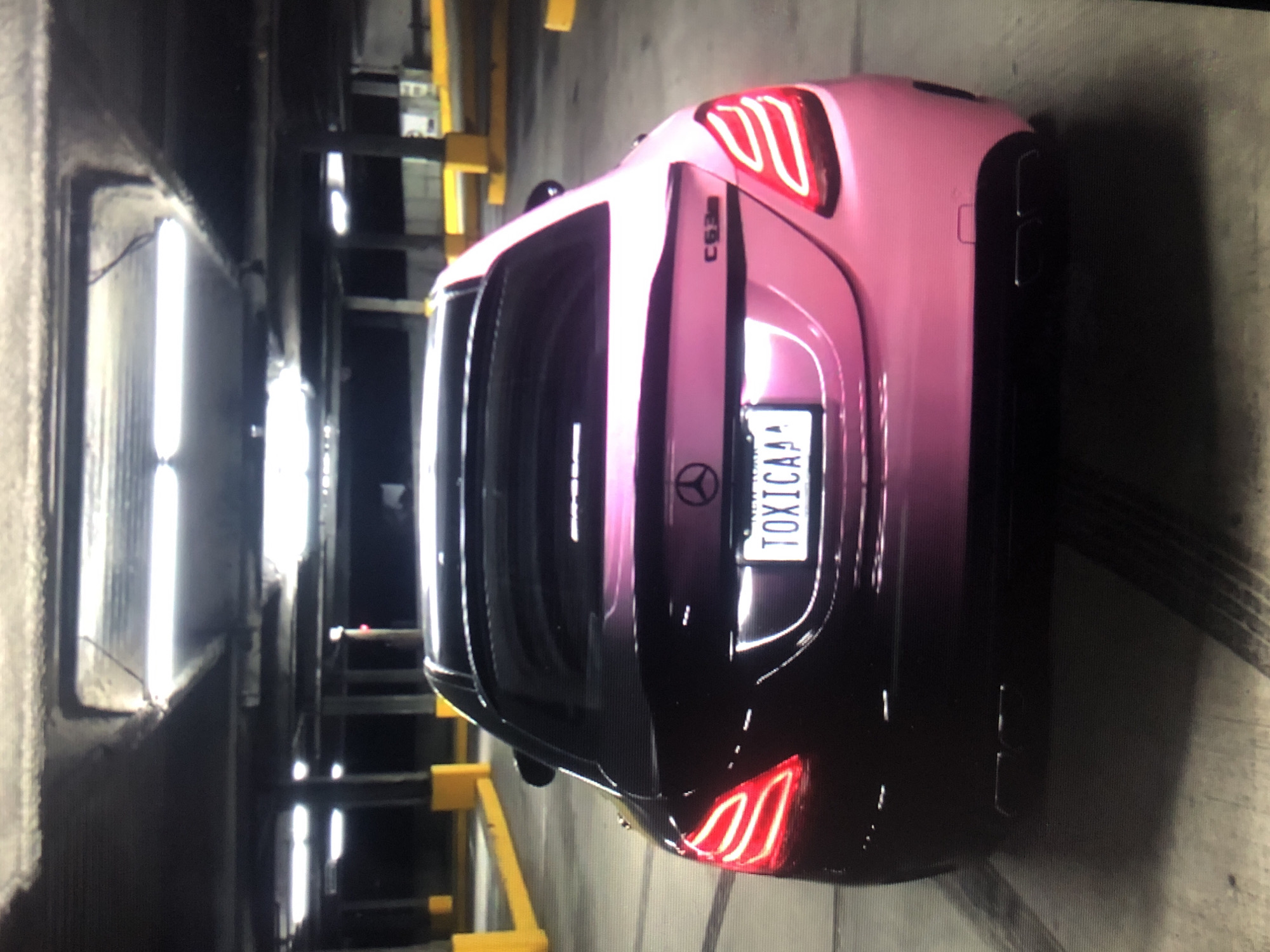 A pink-to-black gradient Mercedes-Benz C63 AMG is parked in an indoor parking structure. The car's rear is facing the camera, illuminated by red taillights. The license plate reads "TOXICAA." The parking structure has concrete walls and floors with metal support beams. Fluorescent lights illuminate the ceiling. The location within Lhokseumawe, Indonesia, cannot be determined from this image.