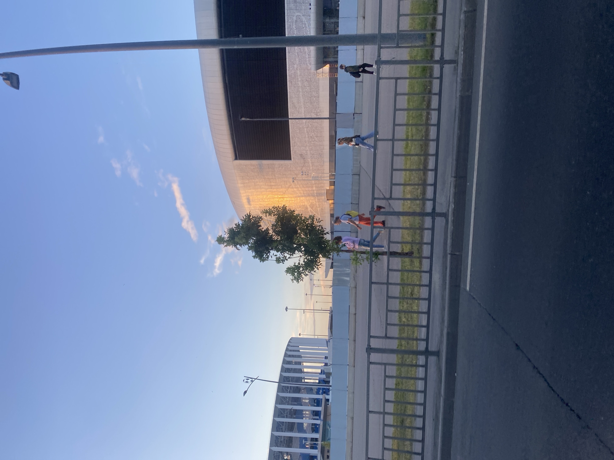 A low-angle shot captures a bright, clear sky over Nizhny Novgorod, Russia. In the distance, a large, modern stadium with a white, curved roof is visible, illuminated by the warm glow of the setting sun. To the left, another stadium with a white, vertical facade is also present. A row of metal railings separates a pedestrian walkway from a grassy area. Several people are walking along the path, some alone and some in small groups. A single tree stands between the walkway and the stadium. A lamppost with a unique football-shaped fixture is visible on the left side of the image. The foreground shows a dark asphalt road with a white line marking.