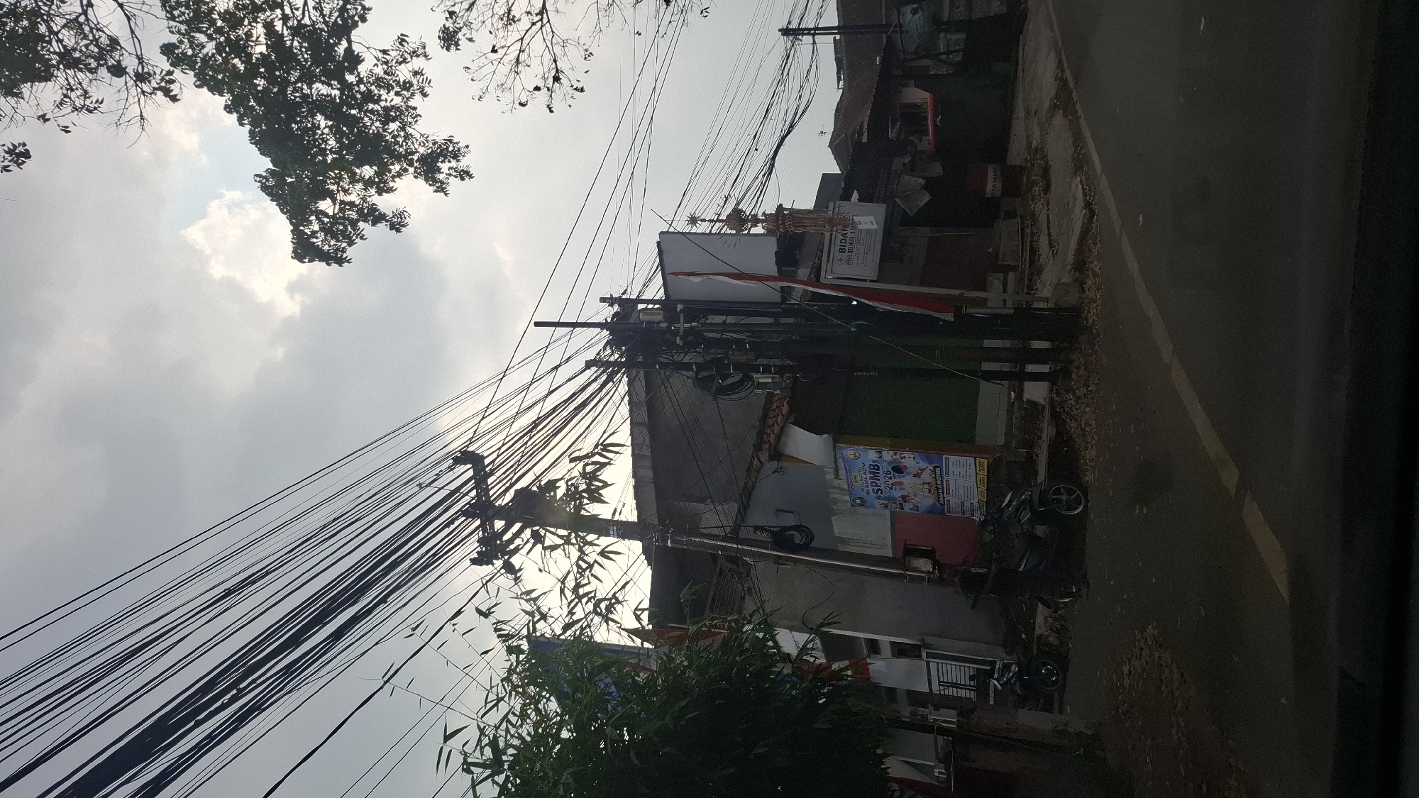 The image depicts a street scene in Cilaku, Indonesia, characterized by a dense network of overhead electrical wires crisscrossing the sky. In the foreground, a paved road with a yellow line marking the lane is visible. To the left, a residential building with a grey facade is partially obscured by lush green foliage. A poster with text in Indonesian, featuring the acronym "SPMB" and the year "2026", is affixed to the building. Two motorcycles are parked on the side of the road. In the middle ground, a utility pole supports the multitude of wires, and a smaller pole with electrical equipment stands nearby. Further down the street, another building with a red and white flag is seen, along with what appears to be a minaret or tower structure. The sky is overcast with diffused sunlight, suggesting either early morning or late afternoon. The overall impression is that of a typical Indonesian urban or semi-urban environment with its characteristic infrastructure and street life.