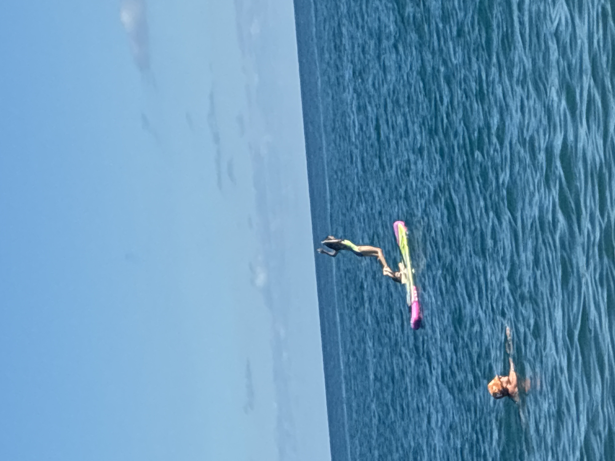 In the image, two individuals are in the ocean under a clear blue sky with scattered clouds. In the foreground, a person wearing an orange hat is swimming. In the background, another person is in the process of jumping off a colorful inflatable mat. The ocean is a deep blue with moderate waves. The location is not identified as Sirius, Russia, as the image depicts a coastal scene.