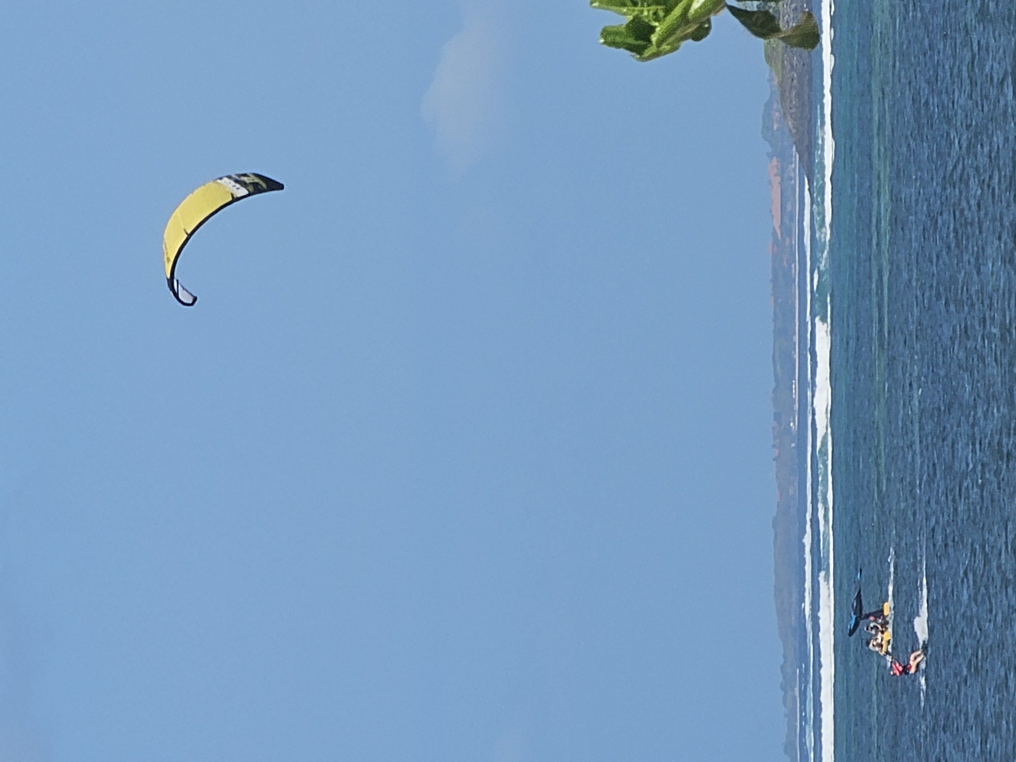 A bright yellow and black kite is suspended in a clear, bright blue sky. Below the kite, on the deep blue ocean, several individuals are engaged in water sports. In the foreground, a person wearing a red life vest is actively kiteboarding, with a spray of water trailing behind them. Further back, a group of people are on a yellow raft-like vessel, with a dark blue object, possibly another kite or sail, attached to it. White waves are breaking in the distance, and a hazy coastline with some vegetation and structures is visible on the horizon. A palm frond is visible in the lower right corner of the image. This scene is likely located off the coast of Denpasar, Indonesia, a popular destination for water activities.