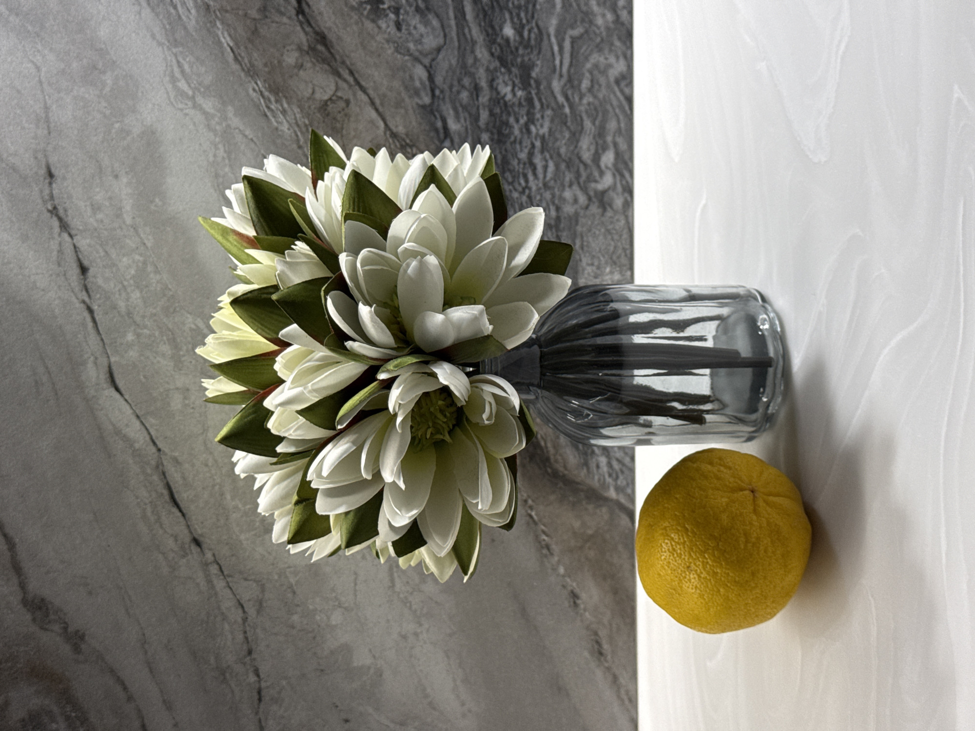 A still life image features a bouquet of white and green artificial flowers in a ribbed glass vase, placed on a white marble countertop. To the left of the vase, a single yellow lemon rests on the countertop. The background is a gray marbled wall. The scene is well-lit, with shadows cast by the objects. There are no people or animals present in the image. The location of this image is not Novosibirsk, Russia.