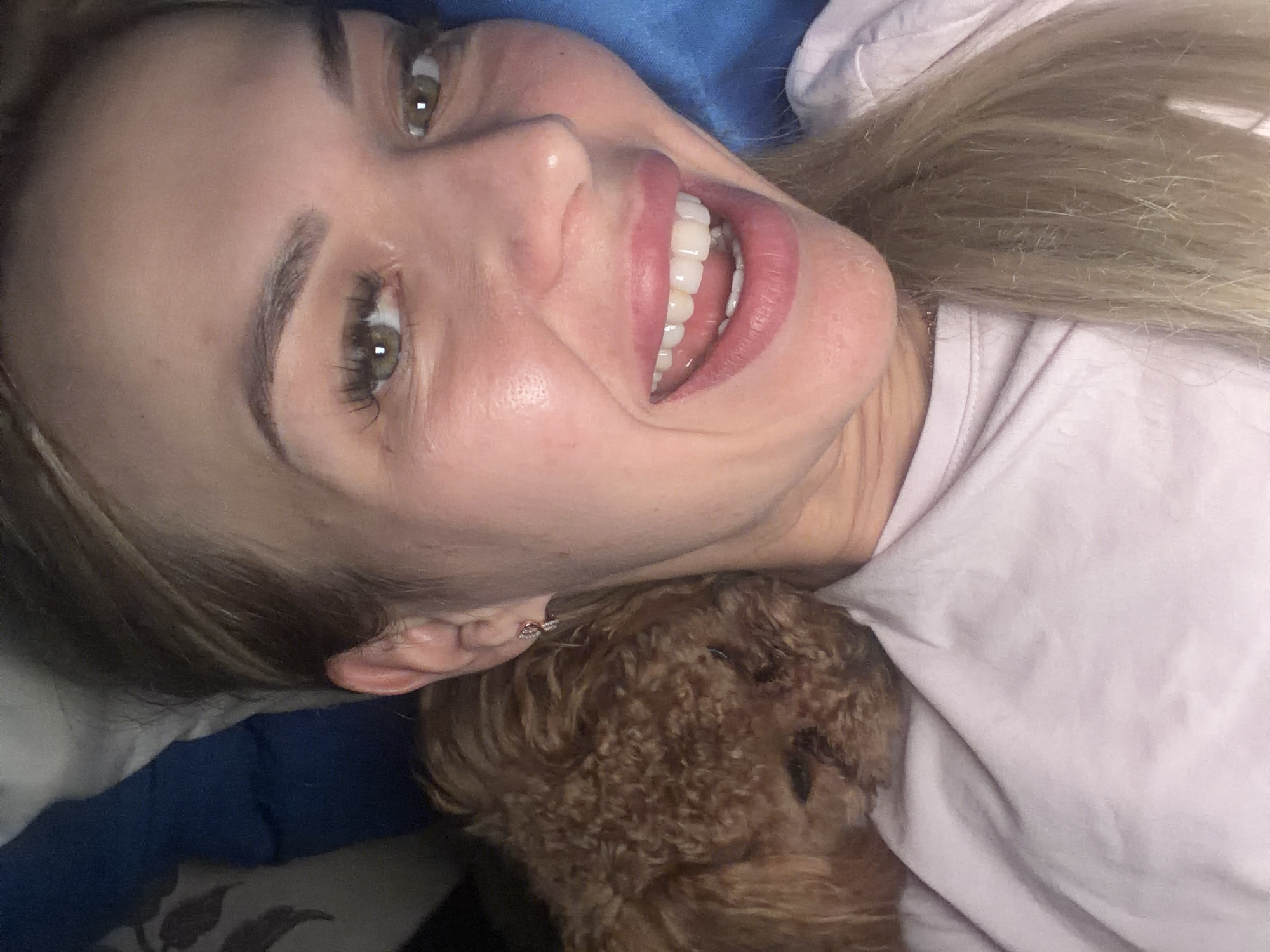 The image is a close-up selfie of a woman and a small brown dog. The woman has blonde hair and is smiling, with her eyes visible. The dog is nestled on her shoulder, with its head resting on her. The background is blurred, but appears to be a bed with blue and white sheets. There is no indication of the location within the city of Kyiv, Ukraine.