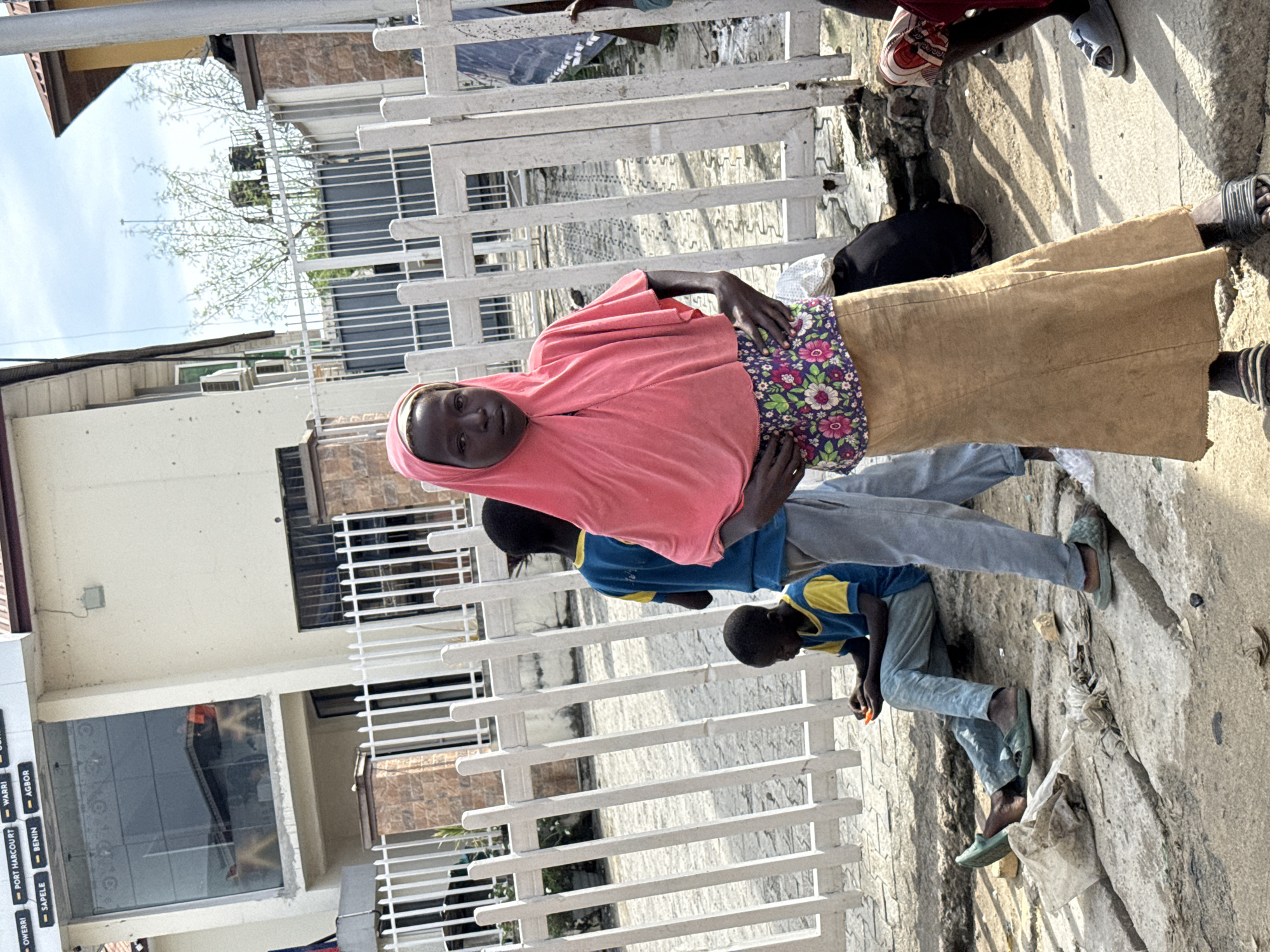 A medium, slightly canted shot captures a group of people standing and interacting in an urban environment, likely Lagos, Nigeria.
The main subject is a woman wearing a pink headscarf, a floral patterned top, and a long tan skirt. She faces the camera with her hands resting on her hips.
Behind her and to her right are two children. One is wearing a blue shirt with yellow sleeves and grey pants, while the other is wearing a blue shirt with a yellow collar and grey pants. They seem to be playing or interacting with something on the ground.
The setting appears to be a street next to a white fence, with buildings visible in the background. The ground is concrete and shows signs of wear and tear. Other individuals are present, indicated by the presence of legs and feet. A sign with the names "Owerri", "Port Harcourt", "Warri", "Sapele", "Benin", "Agbor", and other names is visible on the building behind the fence.