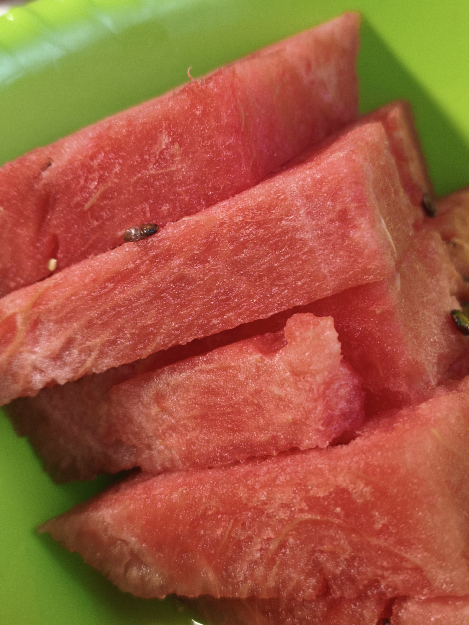 The scene, located in Chaikovsky, Russia, presents a close-up, top-down view of several bright red watermelon slices arranged on a vivid lime green surface. The watermelon pieces are irregularly cut into wedges, displaying their granular, moist flesh and subtle fibrous textures. Multiple small, dark, oblong seeds are visible scattered within the watermelon flesh and occasionally on the cut surfaces. The green background appears to be a plastic plate or shallow dish, with faint reflections indicating a smooth, non-porous material. Shadows cast by the watermelon slices on the green surface suggest a single light source. No individuals or specific urban details of Chaikovsky, Russia, are depicted; the focus is exclusively on the fruit and its immediate container.
