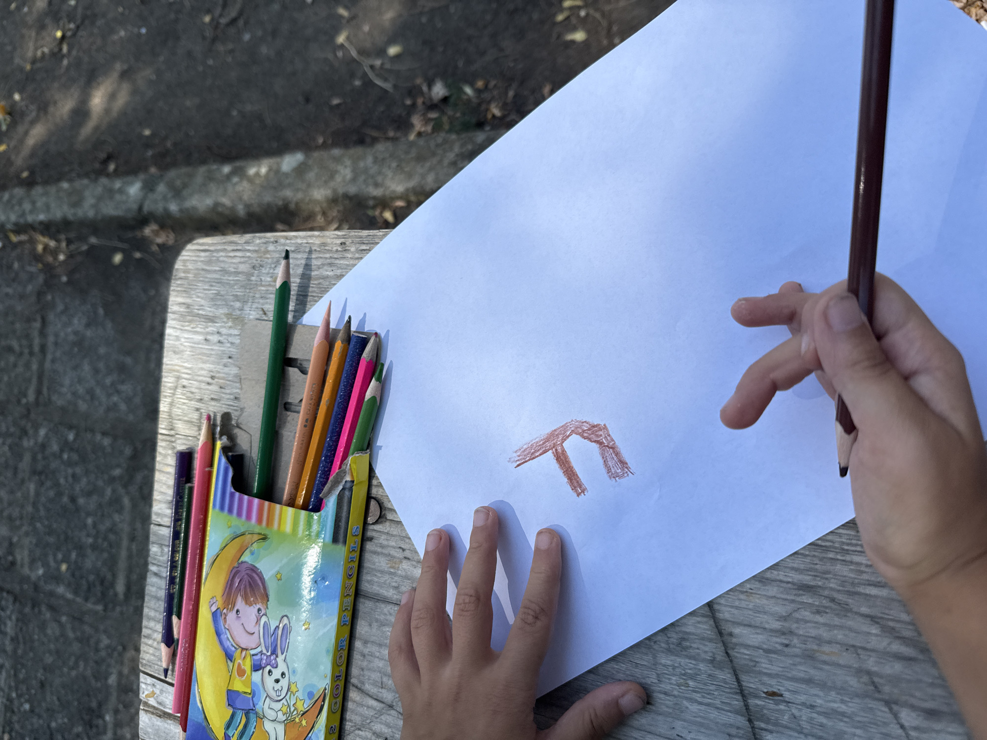 An overhead medium shot shows a child's hands holding a brown colored pencil to a white piece of paper. The child is drawing a brown symbol that resembles a "T" with a hook at the bottom of one of the vertical lines. The child's left hand is resting on the paper, with the fingers spread. The right hand is holding a brown colored pencil and drawing. A box of colored pencils with an illustration of a child with a rabbit is to the left of the paper. The pencils are on a wooden table. The background is blurred and appears to be outdoors with foliage and dirt. The image was taken in the city of Ekzarh Antimovo, Bulgaria.