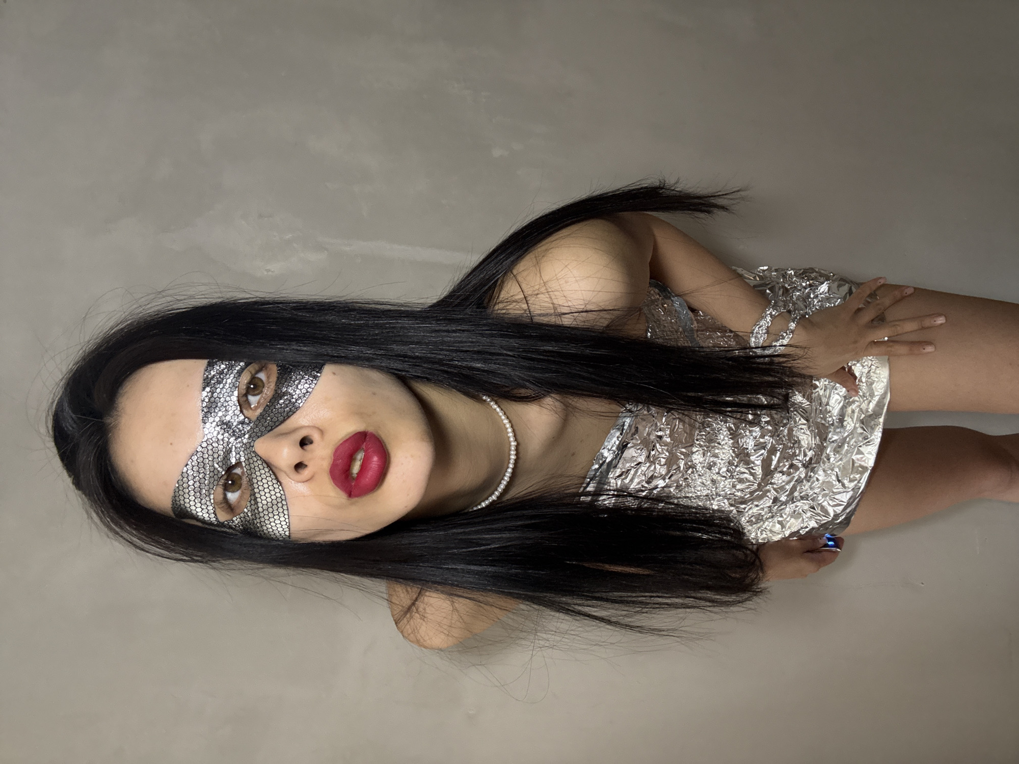A woman with long black hair, red lipstick, and a silver mask is lying on a light gray surface. She is wearing a silver dress that looks like it is made of foil. A pearl necklace adorns her neck. The location is not discernible as Votkinsk, Russia.