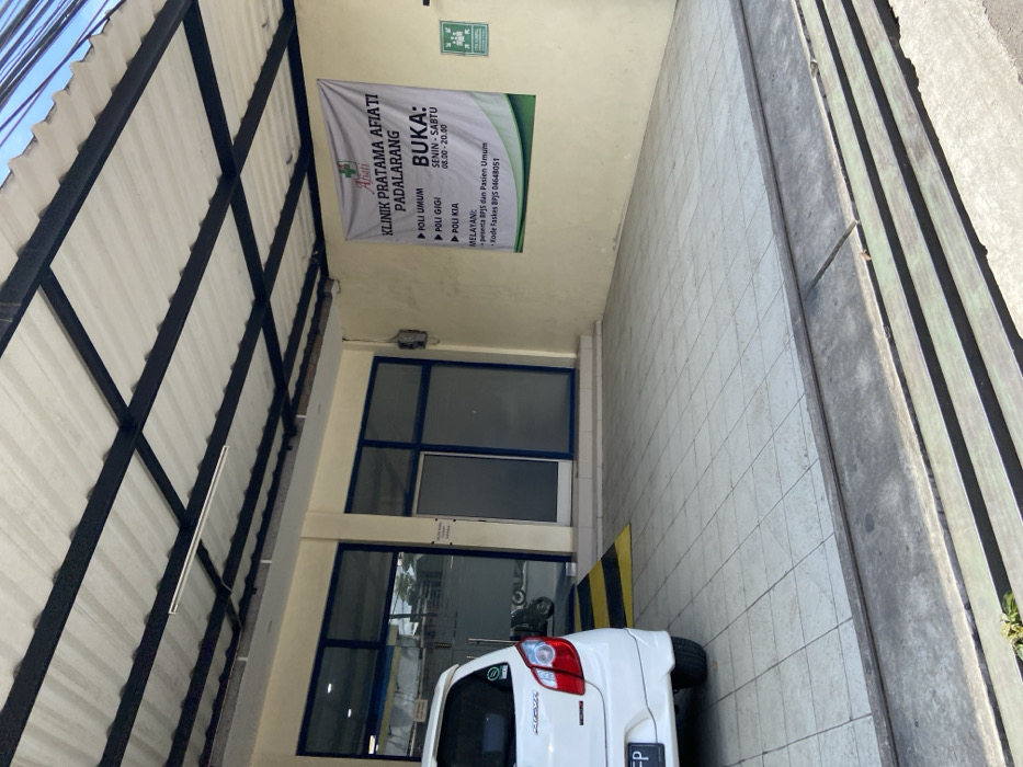 The image shows the exterior of a medical clinic in Padalarang, Indonesia, located at coordinates -6.856 latitude and 107.498 longitude. The clinic is called "Klinik Pratama Afiati Padalarang" and has a sign indicating its services: Poli Umum (General Practice), Poli Gigi (Dental), and Poli KIA (Women and Children). The sign also indicates that the clinic is open from Monday to Saturday, 08:00 - 20:00. There is a white car parked in front of the clinic. The car has a license plate starting with "FP". The clinic has a covered patio area with tiled flooring and steps leading up to the entrance. The building is painted in a light beige color, and the windows are framed in dark blue. The roof is covered in a black material that appears to be metal. The image was taken during the day, as evidenced by the bright lighting and the absence of shadows. There are no people present in the image.
