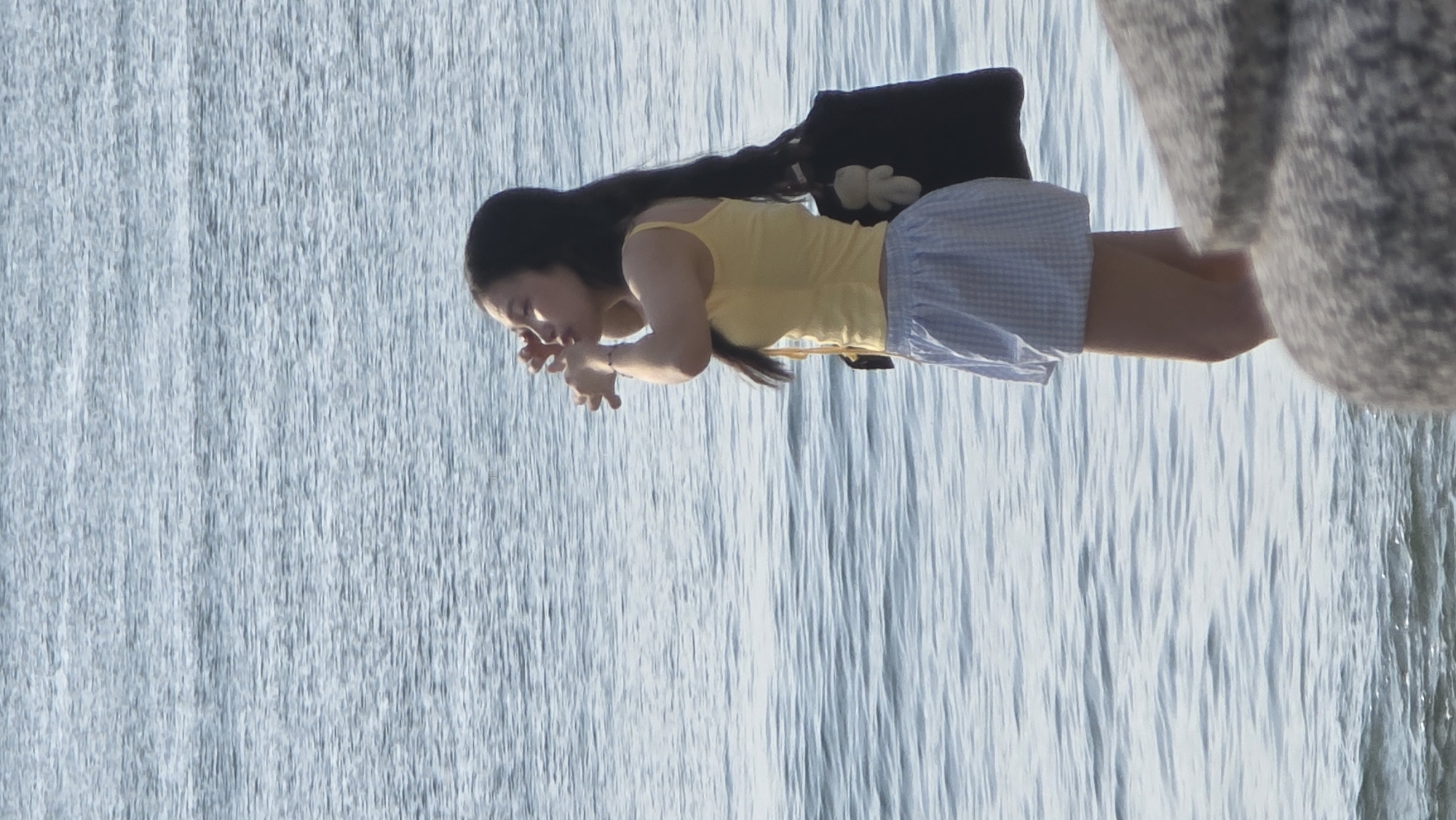 A woman is hunched forward at the edge of a body of water, possibly touching the surface. She has long black hair and is wearing a yellow top and light blue checkered shorts. A black bag is slung over her shoulder. To her right, what appears to be a large grey rock or concrete structure is present. The location appears to be a waterfront, as suggested by the extensive water surface behind her. The scene is outdoors with ample natural light. The image suggests a location in Nha Trang Ward, Vietnam, based on the assignment criteria, but lacks distinct identifying markers of the specific urban environment.