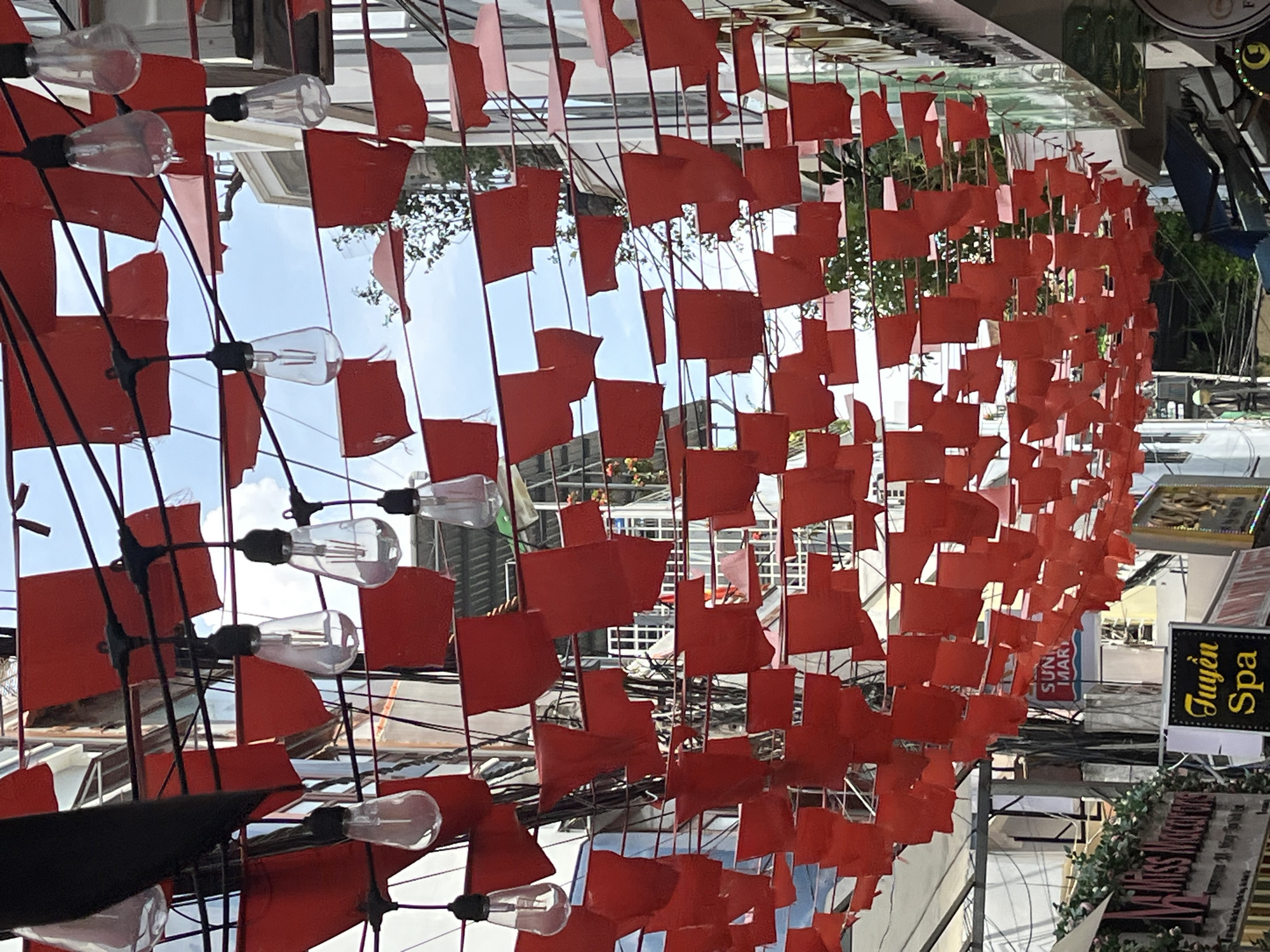 An overhead view captures a street scene adorned with numerous red paper flags and string lights. The flags are strung horizontally across the street, creating a dense canopy. Several clear, Edison-style light bulbs hang from wires interspersed with the flags. The background reveals the architecture of buildings on either side of the street, with balconies and windows visible. The sky above is bright blue with some scattered clouds. At the bottom of the image, signs for businesses are visible, including "Miss World" and "Tuyền Spa," indicating a commercial area. The overall impression is of a festive or decorated street, possibly for an event or as part of the urban landscape in Nha Trang Ward, Vietnam.
