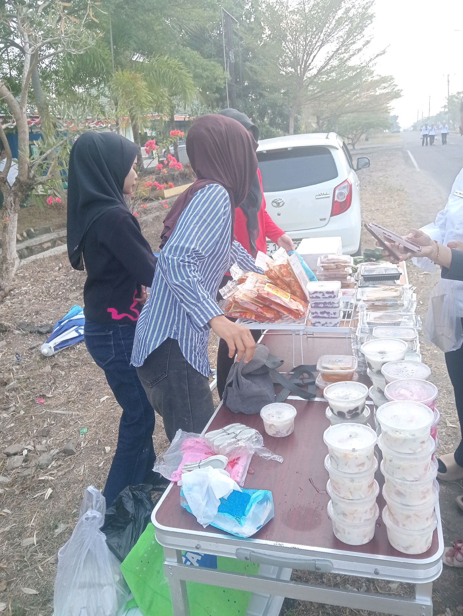 In Sidoharjo, Indonesia, a roadside stall displays various food items in plastic containers on a folding table. Two women wearing hijabs are near the table. One woman on the left, in a black hijab and black jacket, stands with her back to the viewer. The other woman in the center, wearing a maroon hijab and a blue and white striped shirt, is interacting with the food. A white car is parked behind them, and several other people in white shirts are visible in the distance. The area appears to be outdoors, with dry grass and trees surrounding the scene.