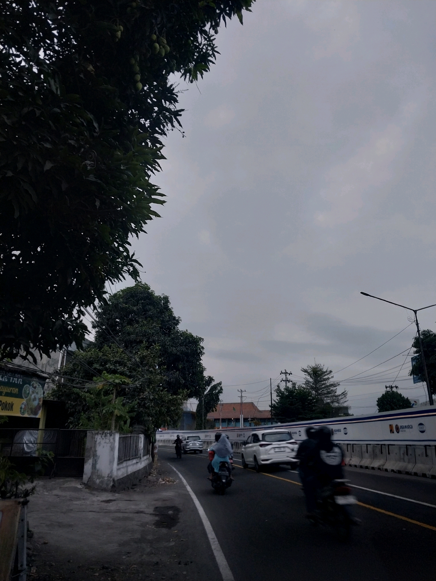 The image depicts a street scene in Maguwoharjo, Indonesia. In the foreground, a dark asphalt road curves to the right. Two motorcycles are traveling on the road; one in the foreground is a dark motorcycle with a rider wearing a dark jacket and helmet, moving away from the camera. Behind it, another motorcycle is also moving away, with a rider wearing a light blue hijab and a light-colored garment. A white car is also present on the road, traveling in the same direction as the motorcycles.
To the left of the road, a sidewalk and a low, weathered white wall are visible. Behind the wall, a dark metal gate leads to a property with some greenery. A sign is partially visible on the left, with Indonesian text. Above the wall, trees with dark green leaves and some visible fruit are prominent. A large tree with dense foliage is also on the left side of the image, extending into the upper left quadrant.
The road is bordered on the right by a concrete barrier topped with a white panel featuring various logos and text, including "JASA MARGA" and "LINTAS." A tall street lamp with a single arm extends from the right side of the road towards the sky. Power lines are visible crisscrossing the sky. The sky is overcast, with a pale gray and cloudy appearance, suggesting either early morning or late afternoon. In the background, trees and buildings are faintly discernible.