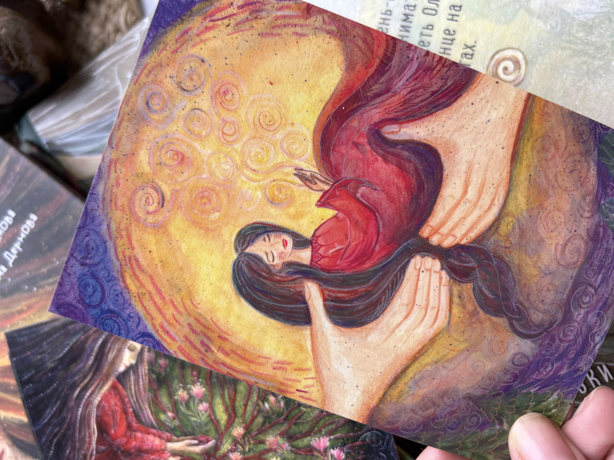 The image shows a close-up of a tarot card depicting a woman lying horizontally within two large, outstretched hands. The woman has dark hair and wears a red garment. A yellow circle with swirling patterns forms a background behind the woman and the hands, transitioning to purple and dark green along the edges of the card. Part of another card is visible in the bottom left corner, showing another woman in a red dress surrounded by what appear to be branches and flowers. There is a third card visible at the top with some unreadable text. The overall style is illustrative and somewhat surreal. The location is not discernible from the image.