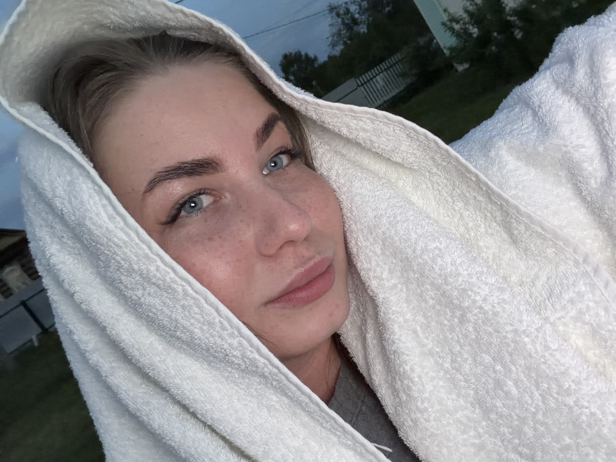 A close-up image shows a fair-skinned woman wrapped in a white towel, possibly after bathing, based on the towel's texture and placement around her head. The towel partially obscures her hair. The woman has light blue eyes, freckles, and shaped eyebrows. The background indicates an outdoor setting, potentially in Уральское, Russia. Visible elements include a fence, trees, and a building, suggesting a residential area. The sky appears overcast or it is near dusk.