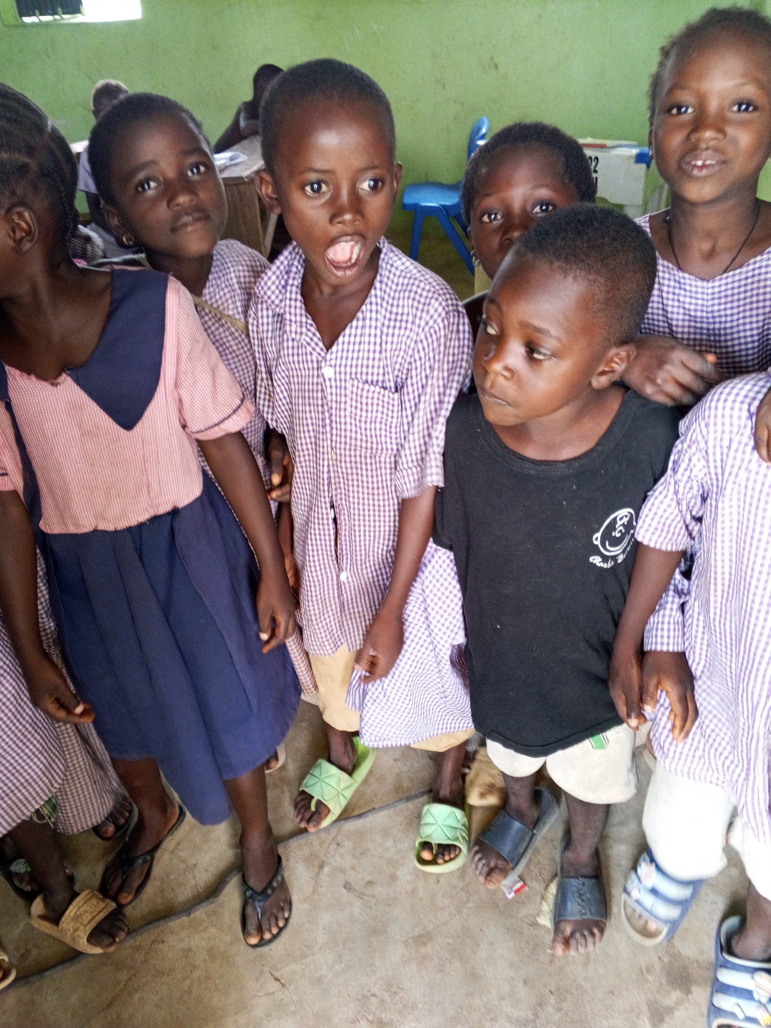 A group of six young Black children, appearing to be students, are clustered together in what seems to be a classroom in Andaha, Nigeria. The child in the front center, wearing a black t-shirt, looks to their left with their mouth open. To their right, a child in a purple and white checkered uniform stares forward. To the left of the child in the black t-shirt, another child in a purple and white checkered uniform with a wide mouth is looking to their right. Partially obscured behind them, a child in a pink and navy blue uniform looks to their left. Two other children are visible in the background, one in a purple and white checkered uniform, and another in the far back wearing a blue and white checkered uniform. The floor is concrete, and a thin, dark cord runs across it. The wall behind the children is painted a light green.