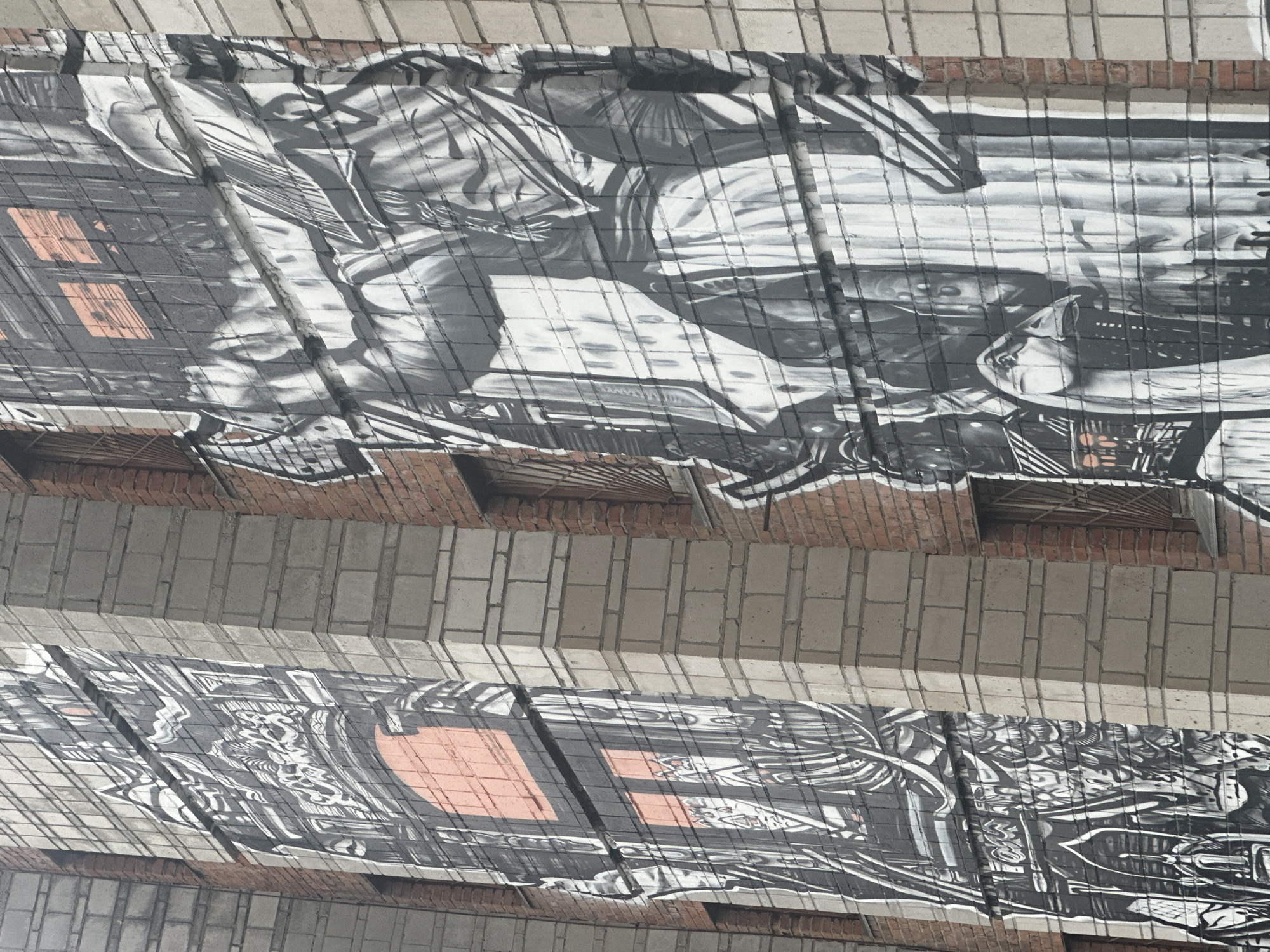 The image shows graffiti on the underside of a bridge or elevated structure in Novosibirsk, Russia. The artwork consists of black and white drawings covering the concrete surface, with the grid pattern of the concrete showing through. The drawings are complex and appear to depict figures and abstract designs. The structure is composed of rows of concrete blocks separated by brickwork. There are gaps and holes present in the brickwork.