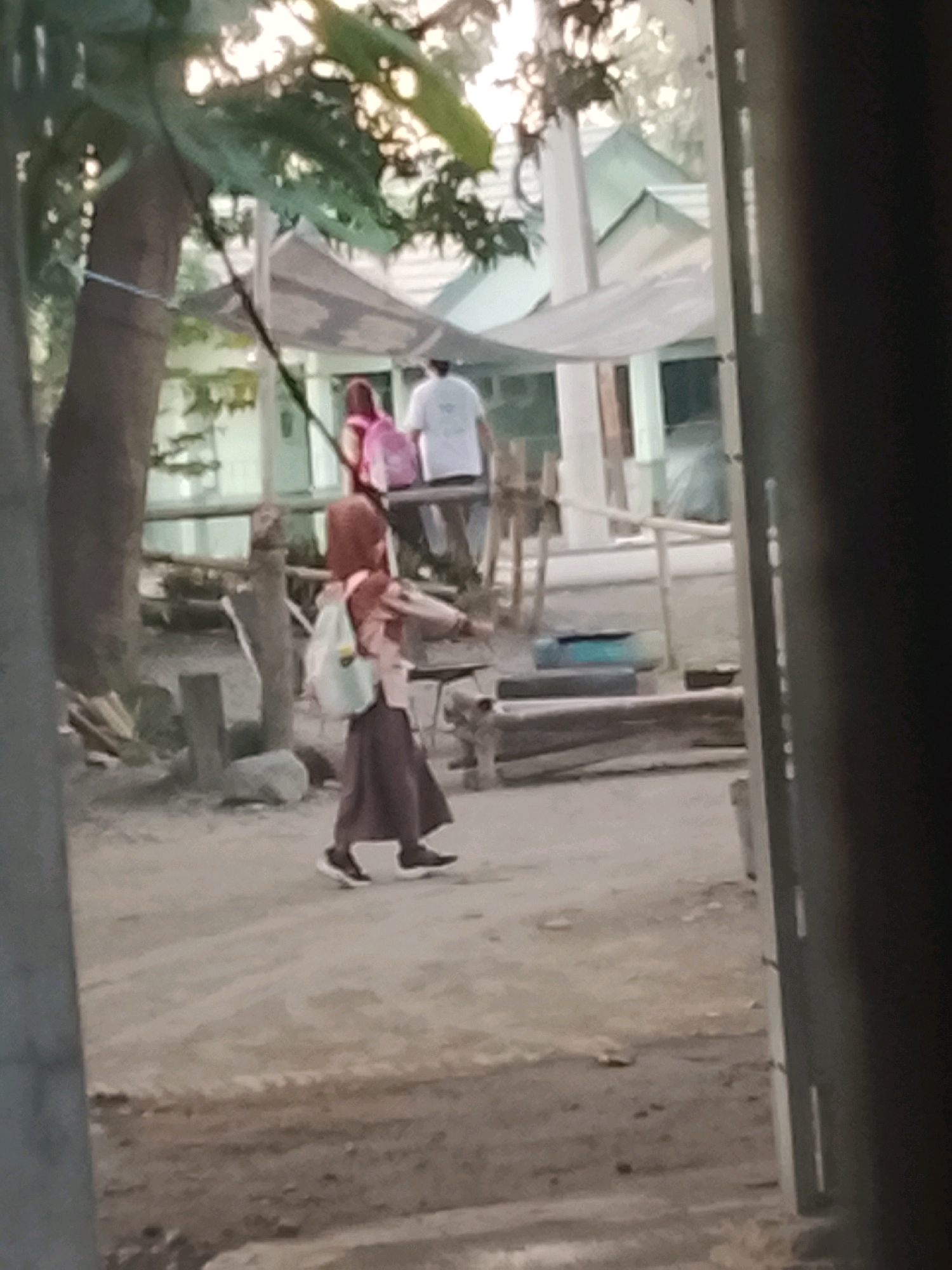 The image, likely taken in Ujungrusi, Indonesia, depicts an outdoor scene with a young girl in the foreground. She is walking from left to right, wearing a hijab and a long skirt, with a backpack slung over her shoulder. To the left, a large tree trunk is visible, and in the background, there are structures resembling buildings with green roofs, possibly part of a school or community area, with awnings providing shade. Other people are present in the background, including one wearing a white shirt and another with a pink backpack. The ground appears to be dirt or sand, with some scattered debris and wooden posts in the midground. The overall lighting suggests it might be late afternoon or early morning.