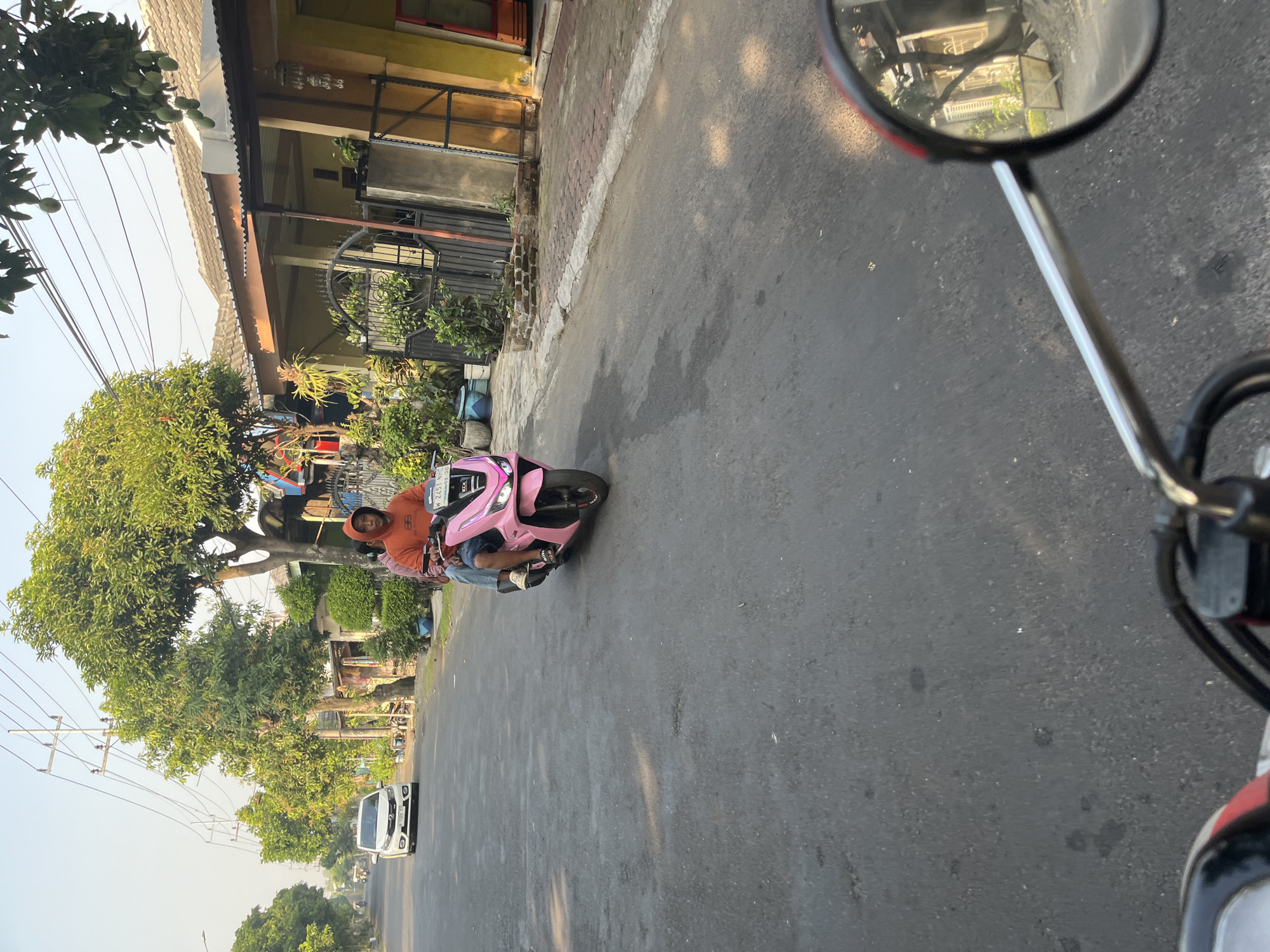 A person wearing an orange hooded sweatshirt and blue jeans rides a pink scooter down a paved street in a residential area. The scooter has a license plate that reads "W 2451 NHP". The person is looking forward, and the scooter is moving away from the camera. In the background, there are houses with green trees and manicured bushes. A white car is parked on the side of the road to the left of the scooter. The sky is clear and bright. This scene appears to be located in Karangtanjung, Indonesia, as indicated by the license plate format and the typical residential architecture and vegetation seen in the image.
