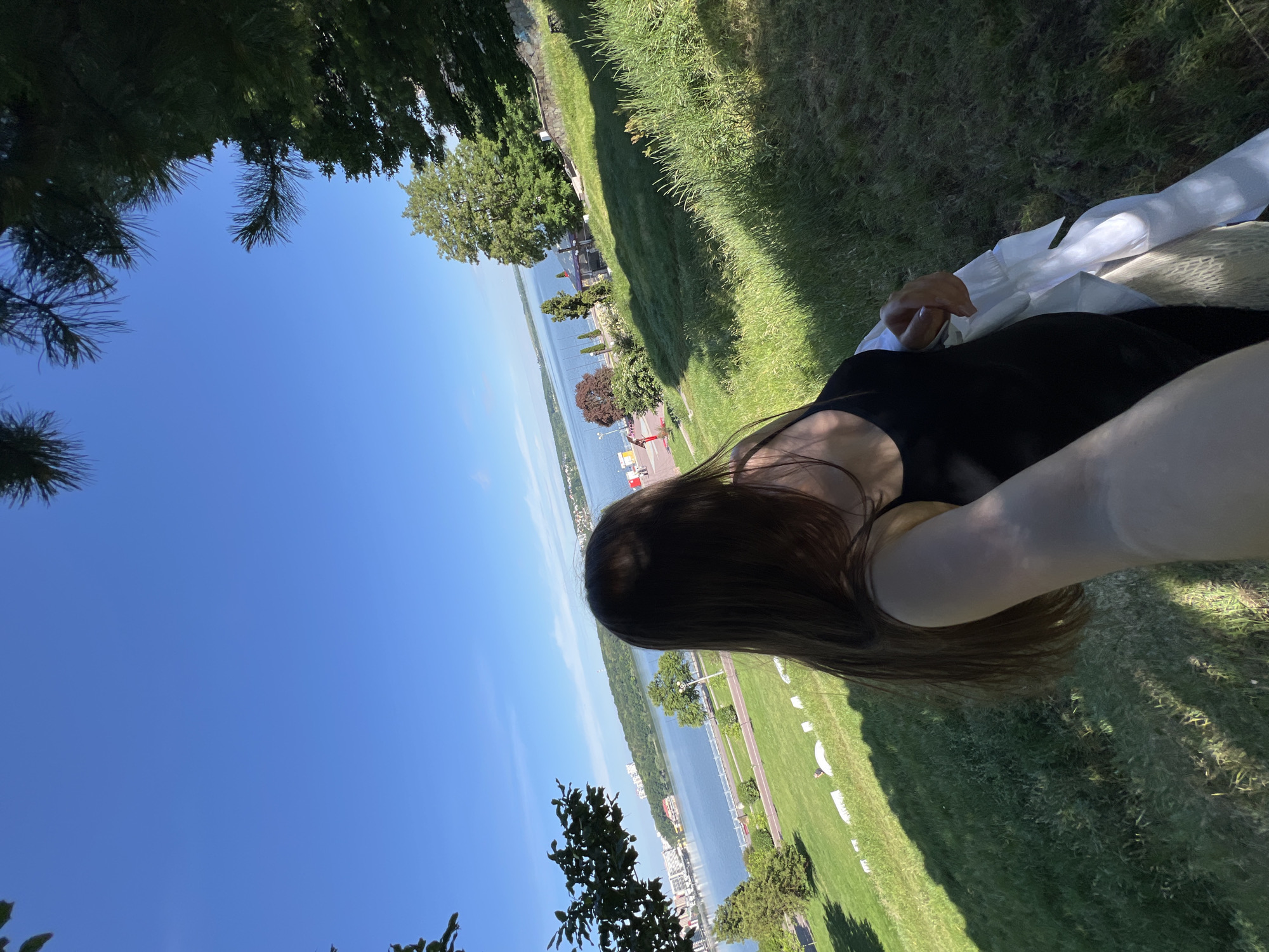 A selfie captures a young woman from behind, her long brown hair obscuring her face. She wears a black tank top and is holding a white shirt. She stands on a grassy hill, looking out at a wide body of water, likely Ternopil Lake, given the city skyline visible on the far shore. Trees frame the scene, with a clear blue sky overhead. The foreground is dominated by lush green grass, dappled with sunlight and shadow. In the distance, buildings and a few scattered trees dot the landscape along the water's edge. The overall impression is one of a peaceful, sunny day in a park setting.