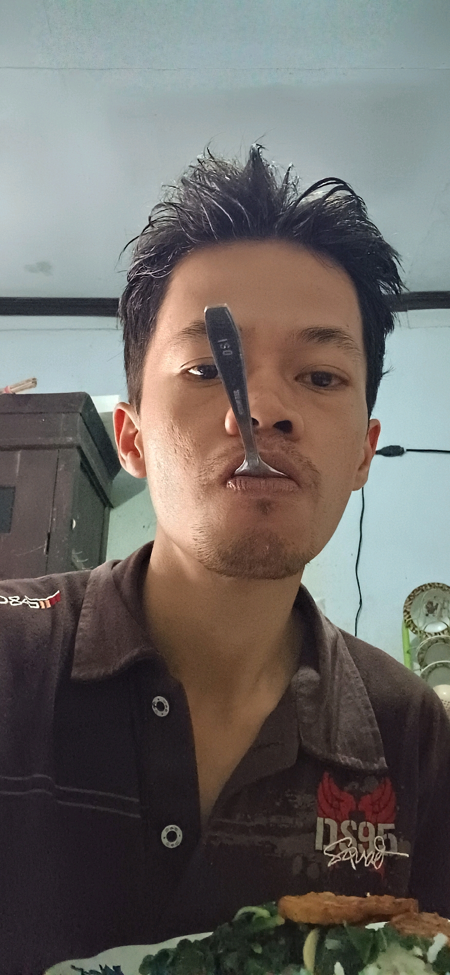 The image shows a close-up, waist-up view of a young man holding a spoon vertically in front of his nose and mouth. He is indoors, as indicated by the wall in the background. He is wearing a dark-colored collared shirt with some emblem or graphic design on the left chest. A plate of food, consisting of green leafy vegetables and what appears to be fried food, is visible at the bottom of the frame. The man has short, dark, and somewhat messy hair. The environment appears to be a home or a simple dwelling. The image is taken from a slightly low angle. The location is specified as Wonokerso, Indonesia.