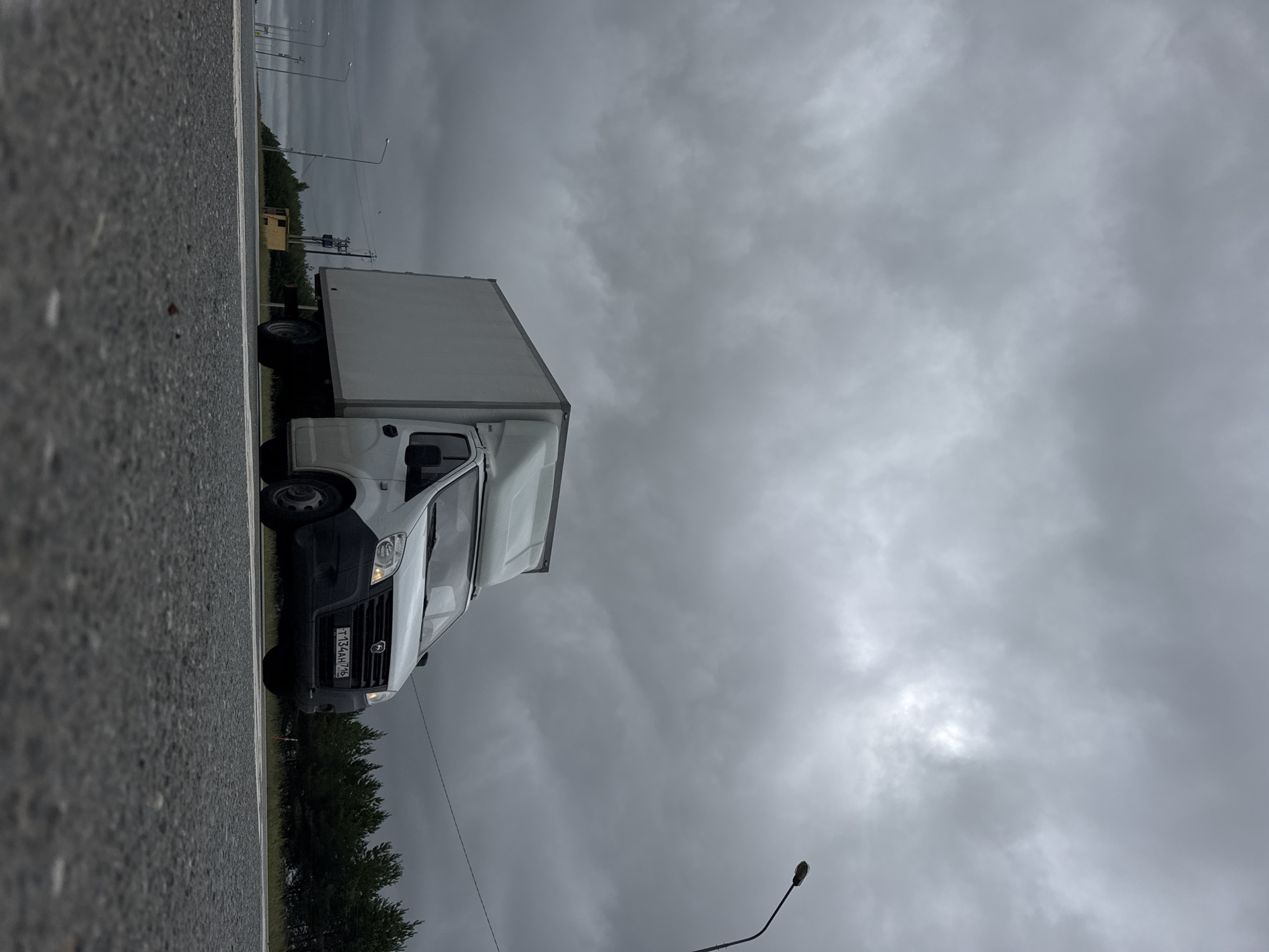 A white box truck is driving on a two-lane asphalt road under a cloudy sky. The truck is in the right lane, and its headlights are on. The road has white lane markings. On the left side of the road, there are streetlights and a line of trees. In the distance, there is a small yellow structure. The license plate of the truck is visible and reads "T134AH 716". The image appears to have been taken in the city of Alnashi, Russia.