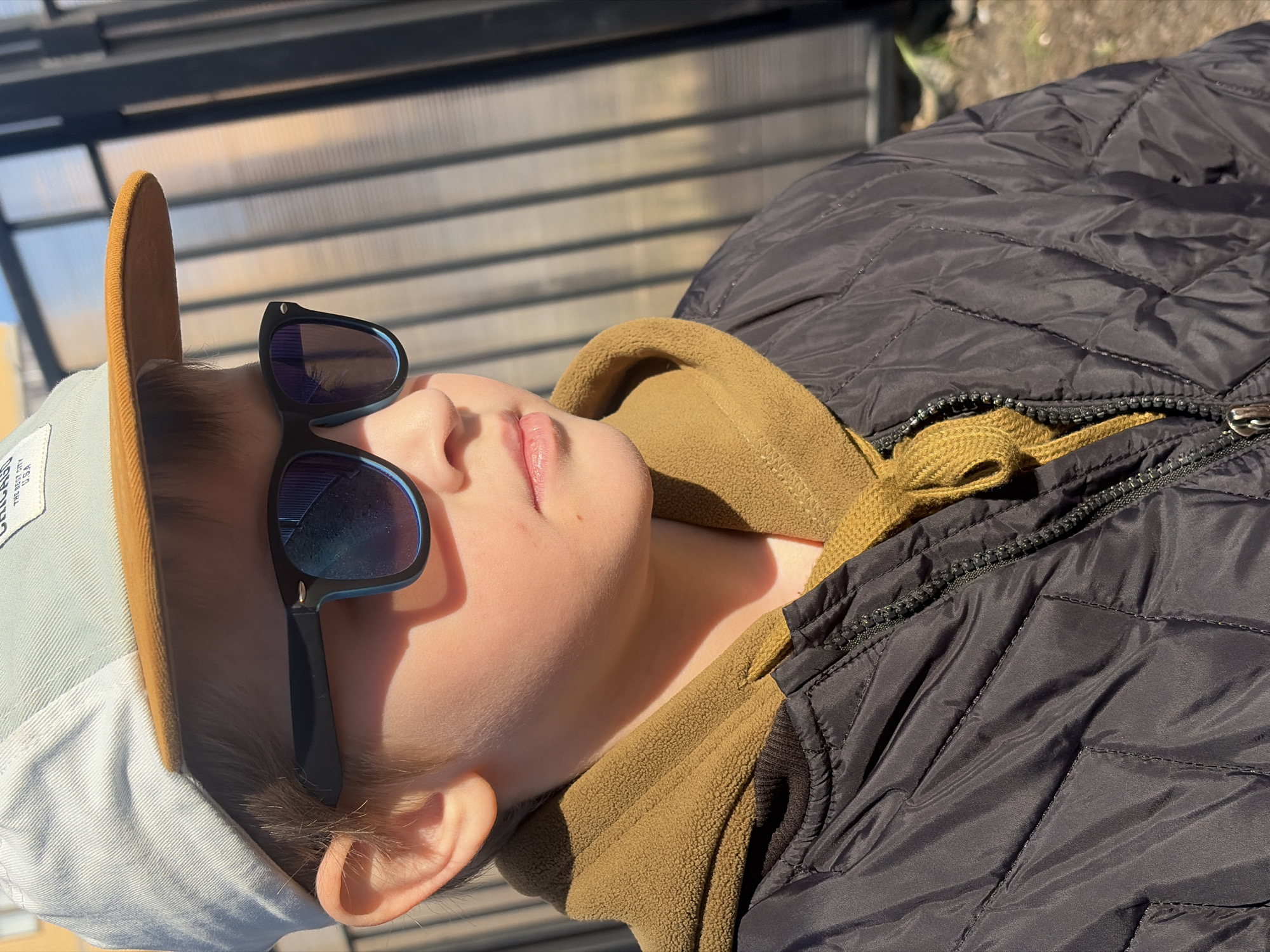 A young person with fair skin is lying on their back wearing a hat and sunglasses, with part of a jacket visible. The hat is light-colored with a brown brim and a white patch with black writing. The sunglasses have dark frames and reflective lenses. The jacket is dark-colored and quilted. The background appears to be outdoors, possibly in Saint Petersburg, Russia, based on the architecture and urban landscape that might be present in the full image, although this is not completely apparent from the cropped view.