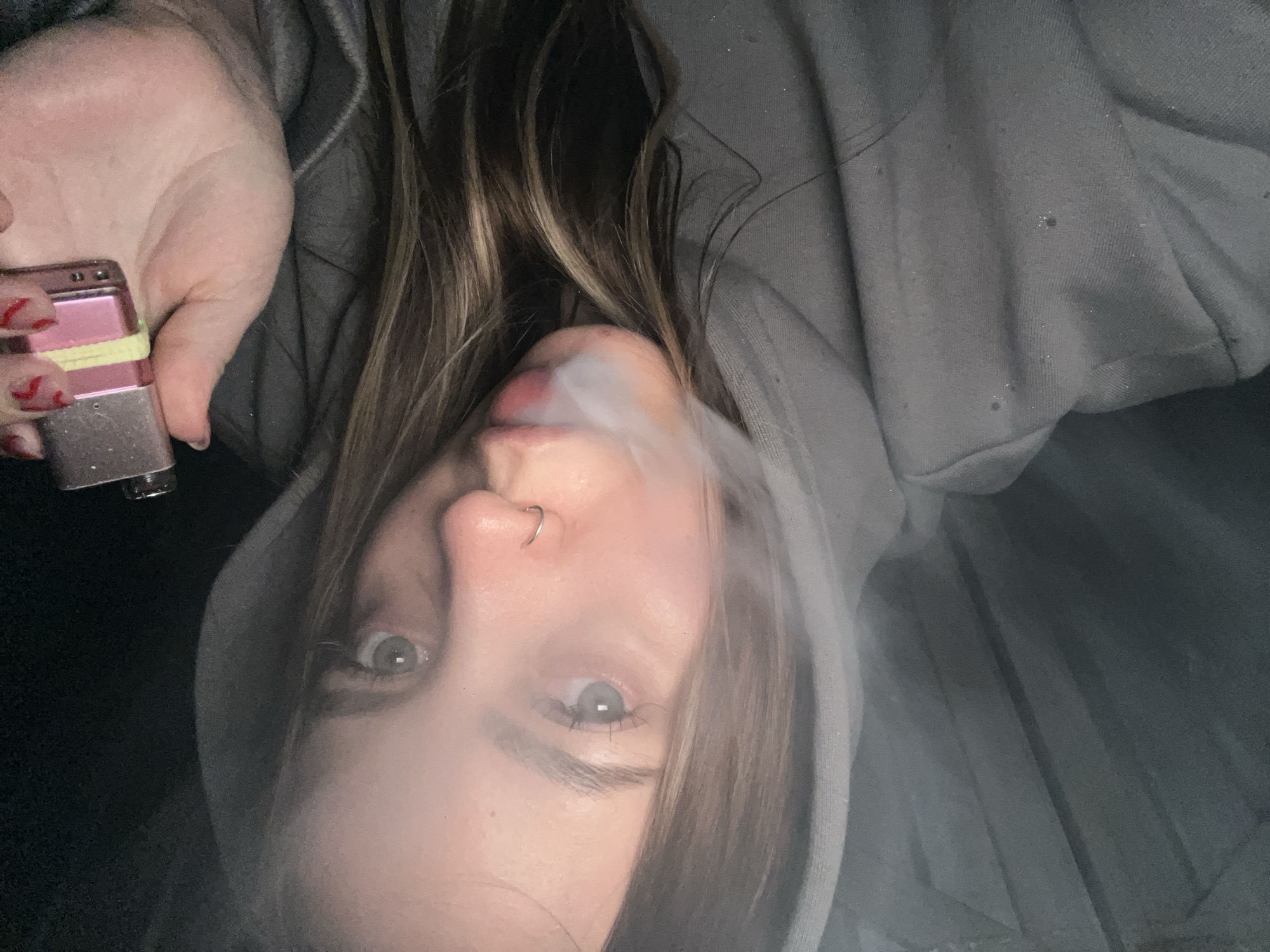A woman in a gray hooded sweatshirt is exhaling smoke from her mouth. She is holding a pink and silver device with a yellow band around it in her hand. Her nails are painted red with black markings. She has a nose ring. No information in the image suggests a location of Novopokrovskaya, Russia.