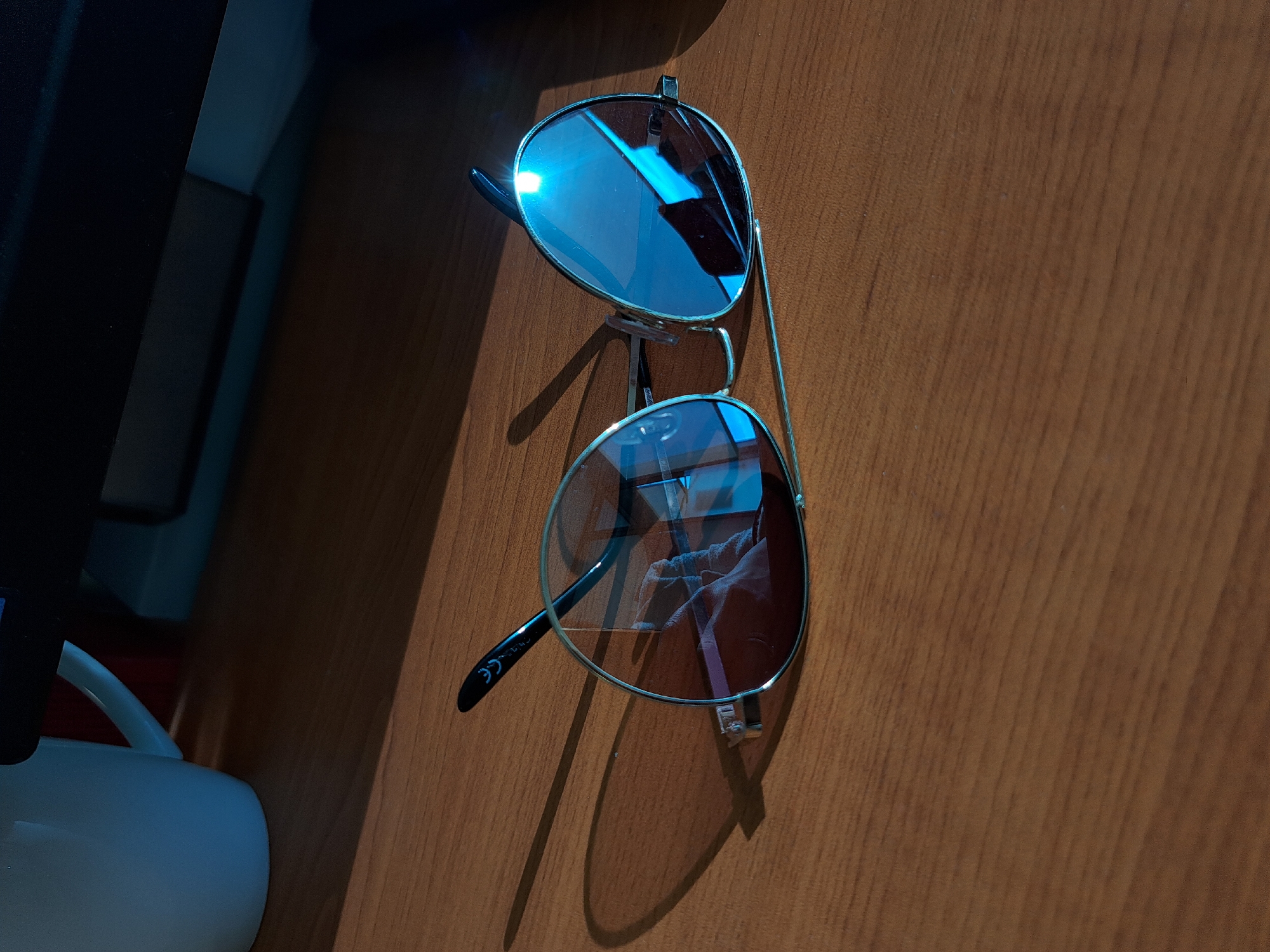 The image shows a pair of aviator-style sunglasses resting on a wooden surface. The sunglasses have a silver frame and blue-tinted lenses. The lenses appear to have some smudges or scratches. A white mug and a computer monitor are partially visible in the background on the left side of the frame. The background features wood grain texture, suggesting a desk or table. The scene is indoors and seems to be illuminated by artificial light. There is no specific information provided to determine that the image location is in Sofia, Bulgaria.