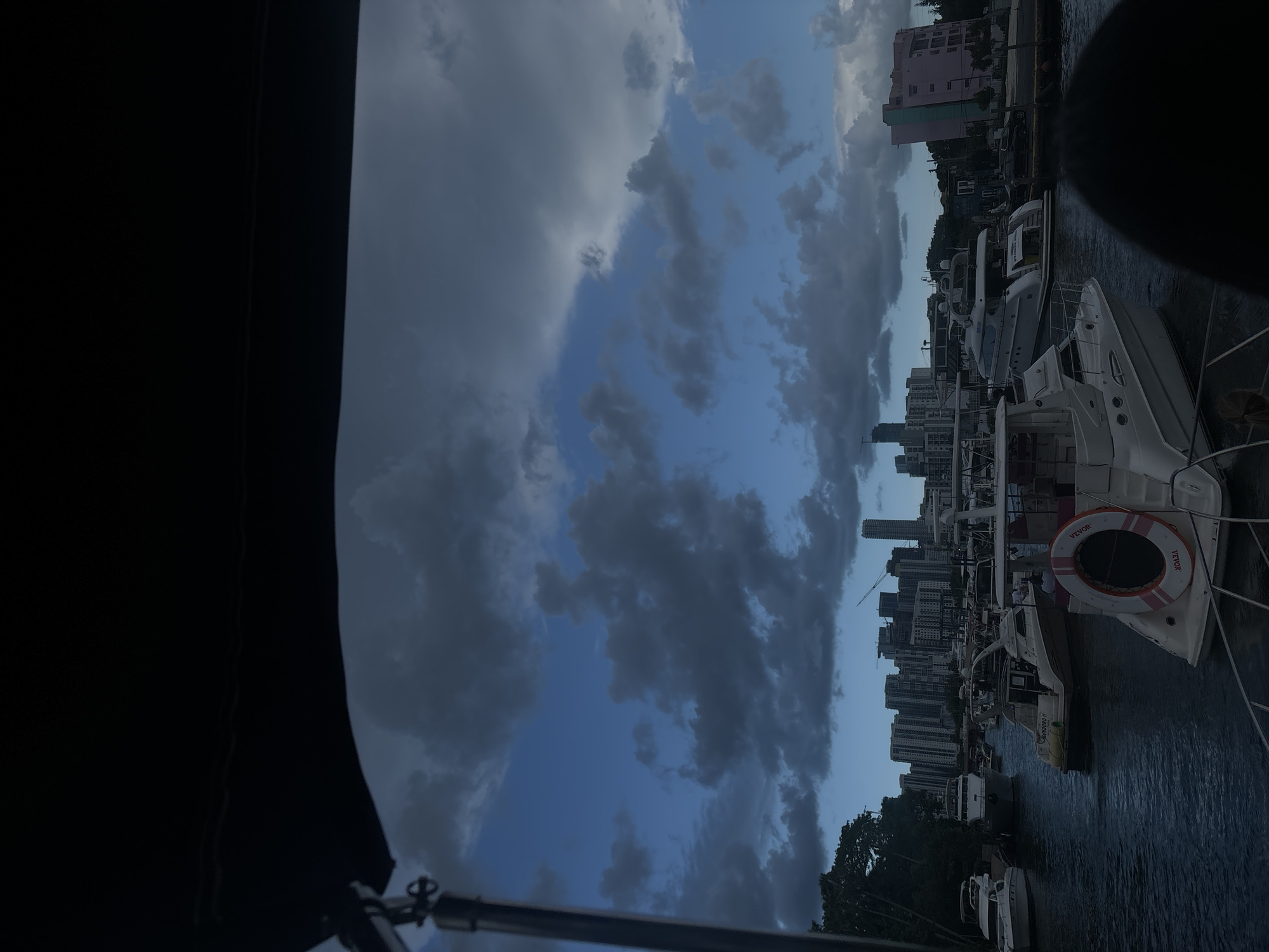 The image depicts a view from a boat in a marina, overlooking a cityscape and the water. The sky is filled with dark, dramatic clouds, suggesting an impending storm or twilight. Several yachts and boats are docked in the water, with a prominent white yacht in the foreground featuring a red and white life preserver. In the background, a dense collection of high-rise buildings, characteristic of a city skyline, is visible. To the right, a brightly colored pink building stands out against the urban landscape. The presence of numerous boats and the distinct urban architecture strongly suggest this location is Miami, Florida.