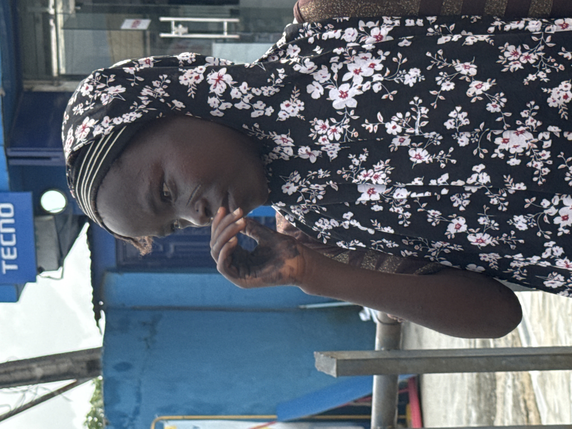 A person wearing a black floral headscarf and matching top stands outside in Ado, Nigeria. The person's face is partially obscured by their hand, and they appear to be looking downward. The environment includes a blue wall, a 'Tecno' sign, and what appears to be a metal railing.