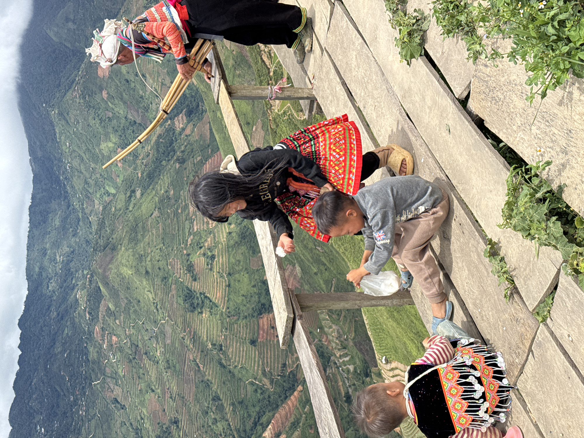 In Ta Xua, Vietnam, on a wooden platform overlooking terraced mountains, two young children and an elder are present. One child, seen from behind, wears a traditional vest with colorful beadwork and fringe, a striped shirt, and jeans. Another child, facing forward, is seated and holding a plastic bag, wearing a grey long-sleeved shirt with a British flag emblem and brown pants. A young girl, in the center, is standing and smiling, wearing a black hoodie and a vibrant red skirt with intricate patterns, and holds a white object in her right hand. To the right, an elder, dressed in a colorful embroidered jacket and black pants, plays a bamboo musical instrument resembling a pan flute. The background features lush, green, terraced hills under a cloudy sky, characteristic of the mountainous landscape of Ta Xua.