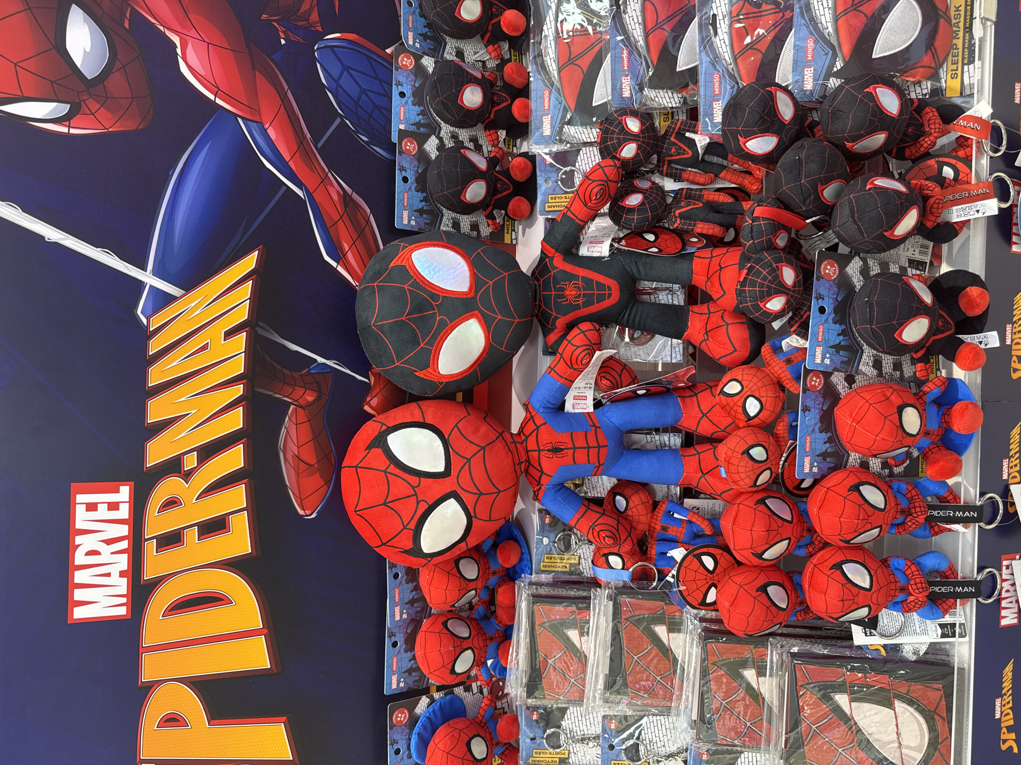 This image depicts a display of Spider-Man merchandise, likely in a retail store. The background features a large graphic with the "MARVEL SPIDER-MAN" logo and an illustration of Spider-Man. In the foreground, there are numerous plush toys of Spider-Man characters in various sizes, including classic Spider-Man and Miles Morales. Some smaller plush toys are designed as keychains. Alongside the plush toys, there are packaged items such as wallets and sleep masks, all featuring Spider-Man designs. The merchandise is neatly arranged on shelves. The location is stated to be Phu Thuy, Vietnam, suggesting this is a retail display in that city. There are no people visible in the image.