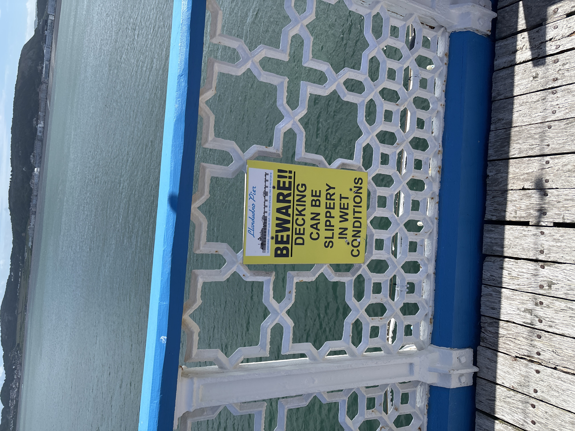 This outdoor shot shows a portion of the Llandudno Pier in Llandudno, United Kingdom. The camera angle is close, focusing on a railing and part of the pier's decking. A yellow warning sign is attached to the railing with the text: "Llandudno Pier BEWARE!! DECKING CAN BE SLIPPERY IN WET CONDITIONS". The pier railing has a geometric pattern in white with accents of blue. To the right, the wooden decking of the pier is visible. Beyond the railing, there is a view of the sea and a distant landmass. The image is slightly rotated clockwise.