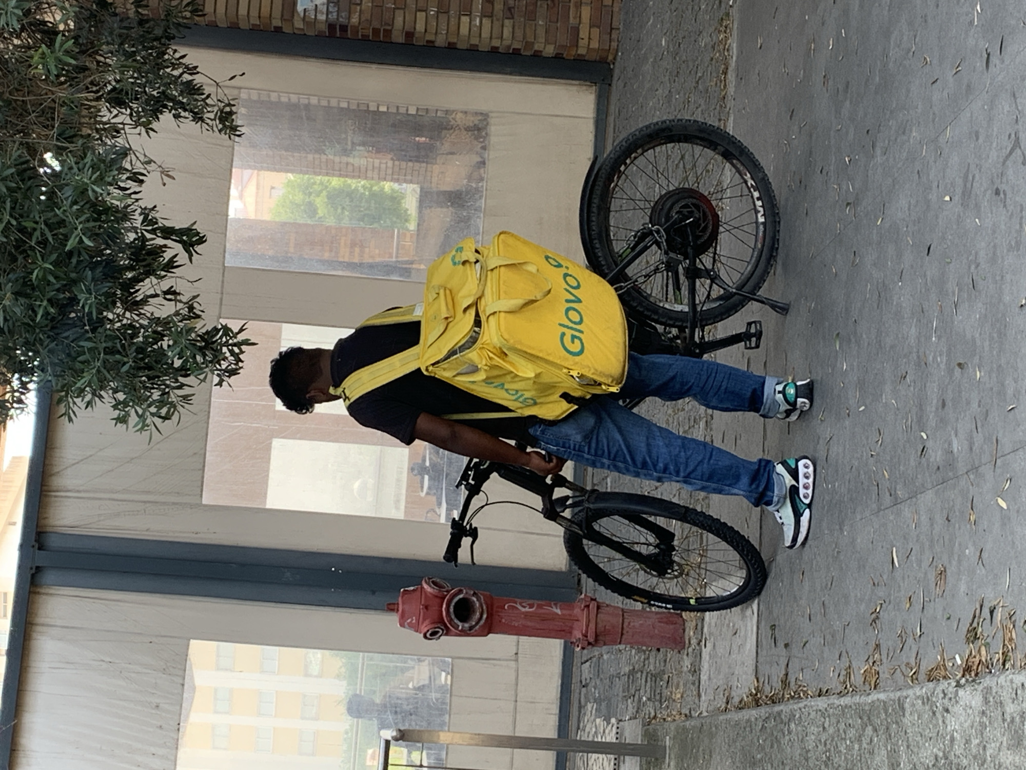 This is a vertical, eye-level shot of a person on a bicycle, possibly a delivery person. The individual is wearing a black t-shirt, blue jeans, and white and green athletic shoes. They have a large, bright yellow delivery bag strapped to their back, with the "Glovo" logo visible on it in green text. The bicycle is a dark color with black tires and appears to be an electric bike. In the foreground, a red fire hydrant is visible. The background consists of a building with large windows and brickwork above. The ground is paved with concrete and scattered with leaves. The scene appears to be outdoors, likely on a street or sidewalk. The lighting suggests it is daytime. The overall impression is a candid, everyday moment of a delivery person on duty.