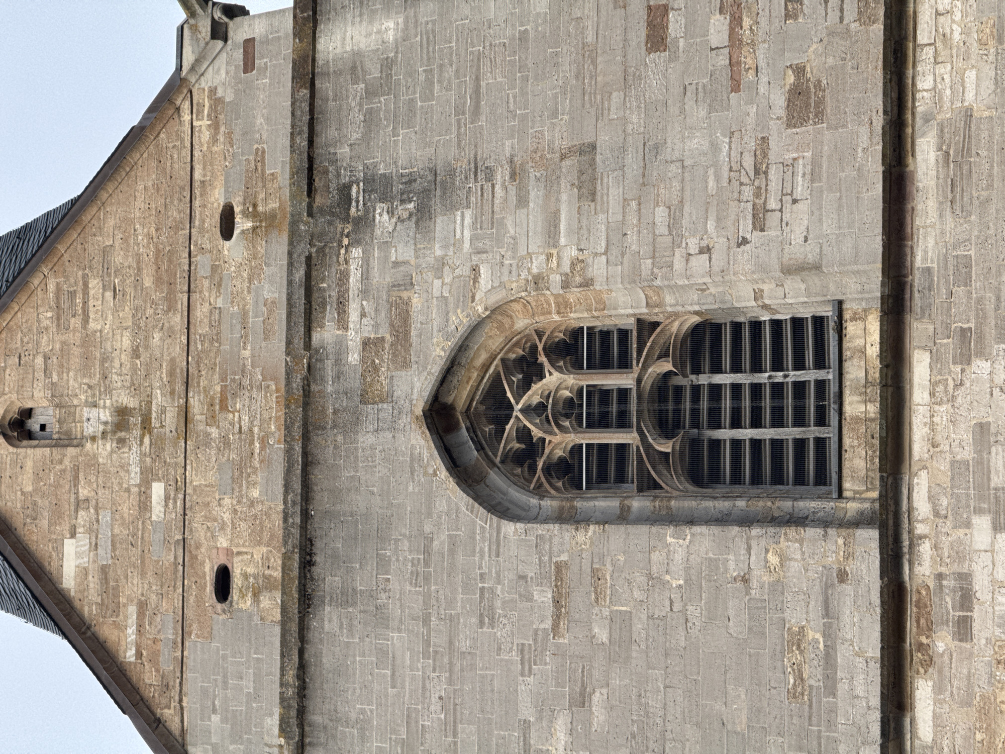 A close-up, low-angle shot captures the facade of a historic stone building, likely a church, with a prominent Gothic arched window. The window is intricately carved with tracery and covered by a metal mesh. The stonework of the building varies in color and texture, ranging from rough-hewn grey blocks to smoother, lighter-colored stones, some showing signs of wear and staining. Two small, oval openings are visible on the upper section of the facade, above the main window. The roofline, with dark grey tiles, slopes down from the peak of the building. The sky above is overcast. The scene is devoid of people or any visible activity.