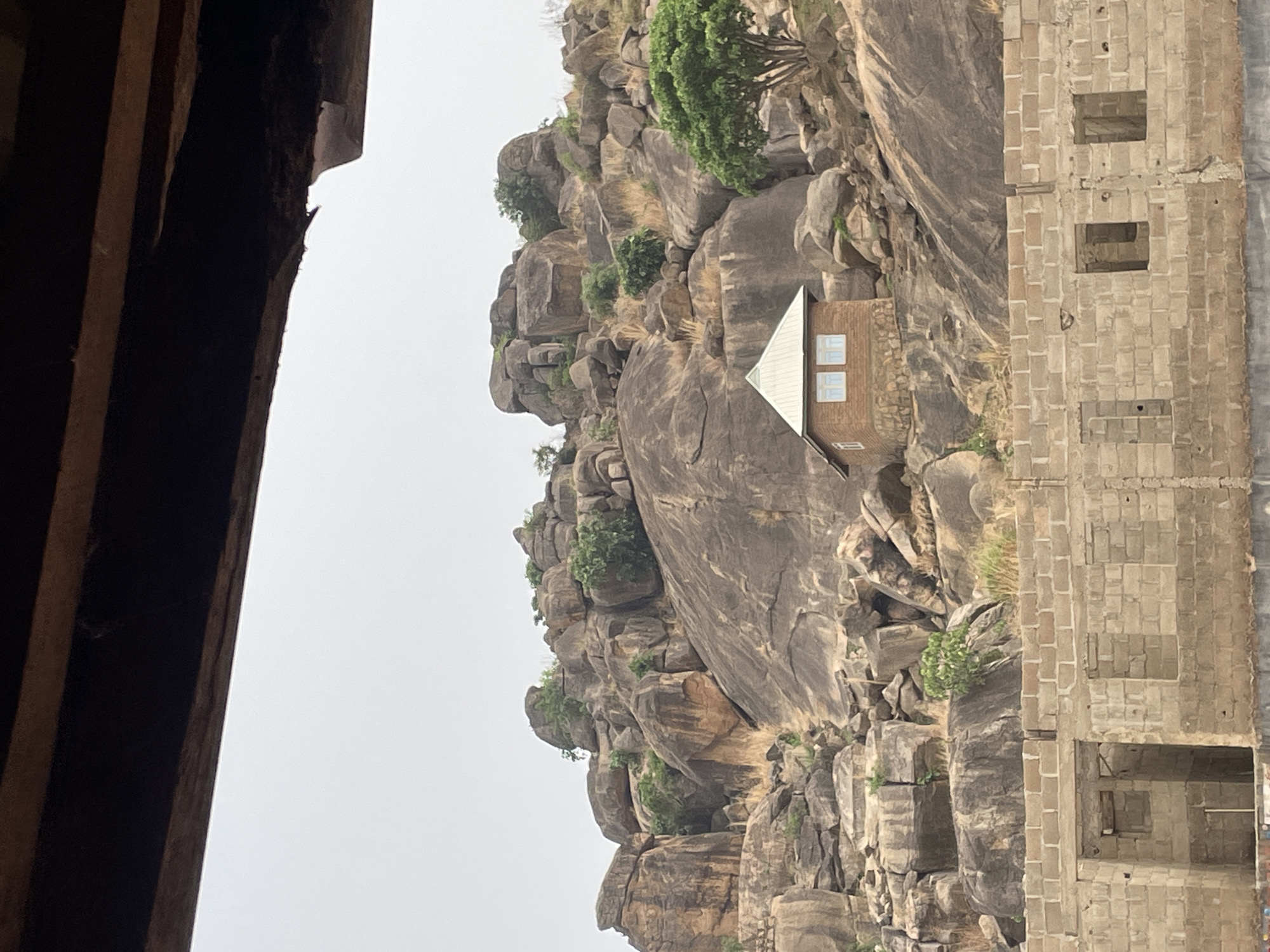 The image shows a rocky escarpment at Jos, Nigeria. The escarpment contains sparse vegetation and a small brick building with a tin roof is nestled amidst the rocks. Adjacent to the escarpment is a multi-story building under construction, consisting of exposed brick. The foreground of the image is shadowed by a window frame, suggesting the photo was taken from an interior location. The sky is overcast. No people are visible.
