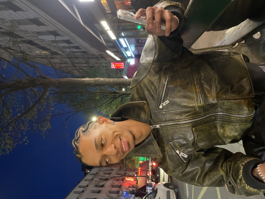 The image depicts a young man on a commercial street in Paris, France, at night. He is positioned centrally, smiling with his eyes directed upwards and to the viewer's left. His hair is styled in close-cropped braids, and he wears an earring in his left ear. He is dressed in a distressed, olive-green leather bomber jacket with a wide collar, a prominent central zipper, and two zippered chest pockets. The zipper pulls on these pockets read "GCDS" and "2002". A silver-toned link bracelet is visible on his left wrist. In his right hand, he holds a transparent smartphone. He appears to be leaning against a dark, possibly metal, barrier.
The background features a tree with sparse leaves and classic Parisian Haussmannian buildings. To the left, a white vehicle is visible on the street. Various illuminated commercial signs, including a red neon sign and a partially visible "MARCHE" sign, line the street, confirming an urban commercial setting. The artificial lighting from streetlights and businesses illuminates the scene against the dark blue night sky.
