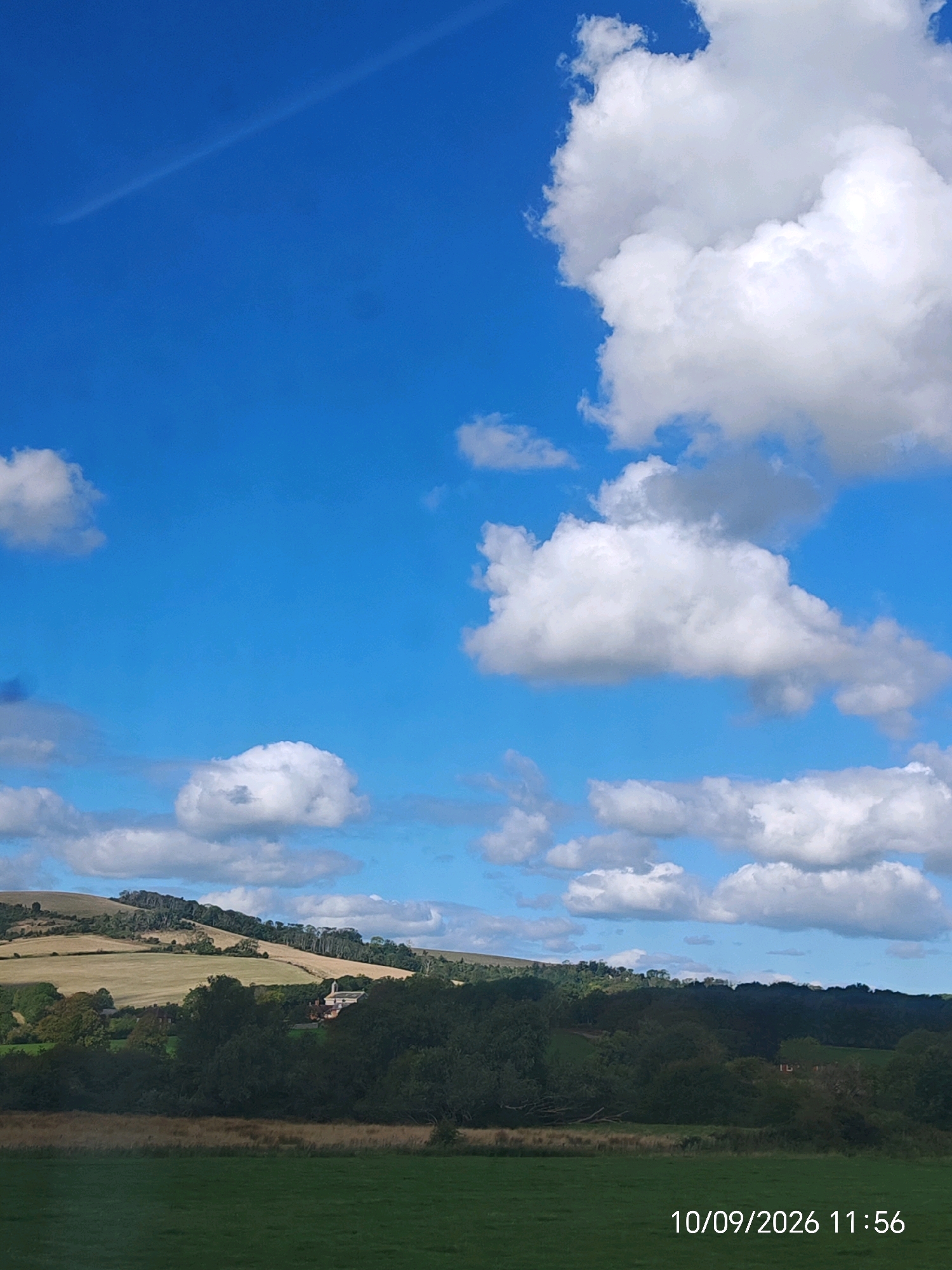 The image captures a rural landscape in Lewes, United Kingdom, under a bright blue sky dotted with fluffy white clouds. A thin, wispy contrail stretches diagonally across the upper left portion of the sky. In the middle ground, rolling hills are covered in a patchwork of fields, some golden brown and others green, with a dense line of trees bordering the lower slopes. A small building, possibly a house or church with a reddish-brown roof, is visible nestled amongst the trees. The foreground consists of a vibrant green field, leading towards the treeline. The bottom right corner of the image displays a date and time stamp: "10/09/2026 11:56".
