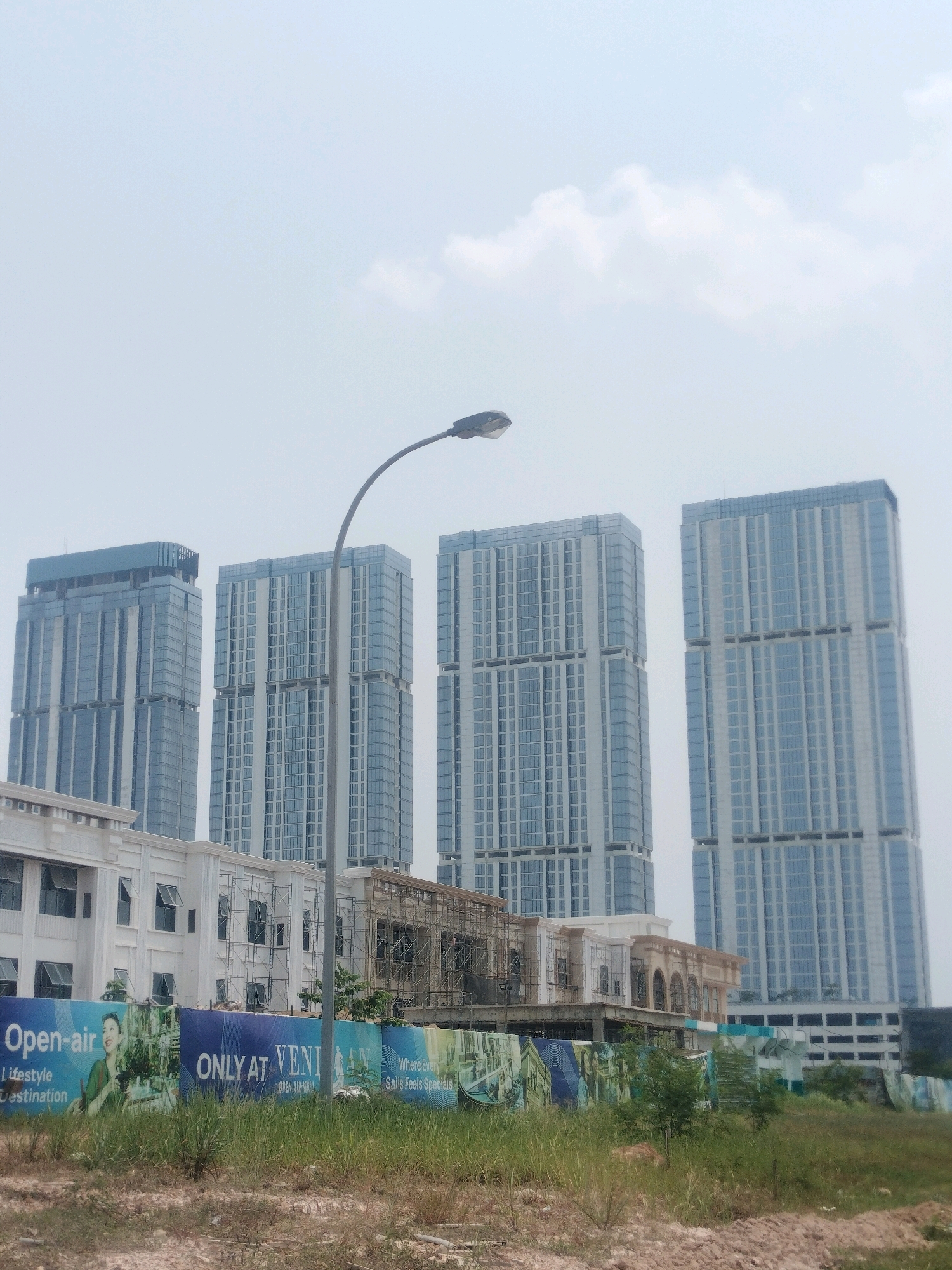 The image shows a construction site in Batam, Indonesia, with several modern high-rise buildings in the background and a lower building under construction in the foreground. A large blue banner with promotional text and images, including the words "Open-air" and "ONLY AT VENI," is visible along the front of the construction site. The sky is overcast with a few clouds. A street lamp curves into the frame from the top left. The ground is covered with dry grass and some soil. The overall scene depicts an urban development project in progress.