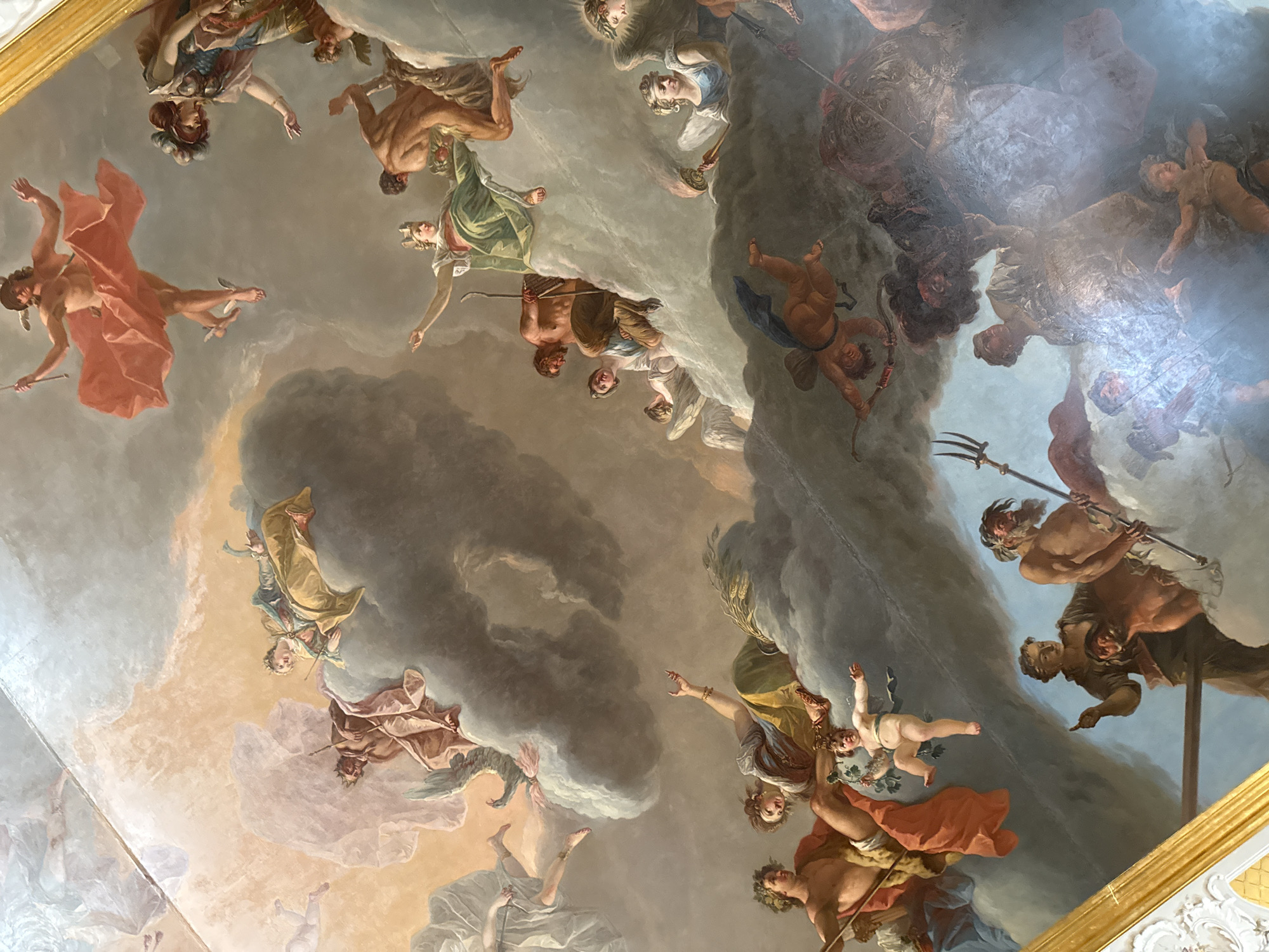This is a detailed fresco on the ceiling of the Grand Hall of the Catherine Palace in Pushkin, Russia. The fresco depicts a grand mythological scene with numerous figures in mid-air, surrounded by clouds and rendered in vibrant colors. The overall composition is dynamic and chaotic, with figures in various poses of movement and interaction.
In the upper left, a figure in a red cloth is depicted as if flying, with wings on its feet. To the right and slightly lower, a woman in a golden robe is shown, possibly an allegory, looking upwards. Below her, a man with a beard and crown is seated, with a figure draped in green cloth beside him.
In the lower half of the fresco, more figures are visible. A bearded man with a trident stands on the left, accompanied by a woman with a laurel wreath and a child reaching out. To their right, a group of figures is engaged in a struggle, with one holding a sword and another appearing to be falling. Several winged cherubs are interspersed throughout the scene, adding to the celestial atmosphere.
The fresco uses rich colors, with dominant tones of pink, gold, blue, and brown, creating a sense of grandeur and drama. The painting's detailed rendering of musculature, drapery, and facial expressions conveys a sense of lively action and emotional intensity. The overall impression is one of a divine or mythological event unfolding in the heavens.