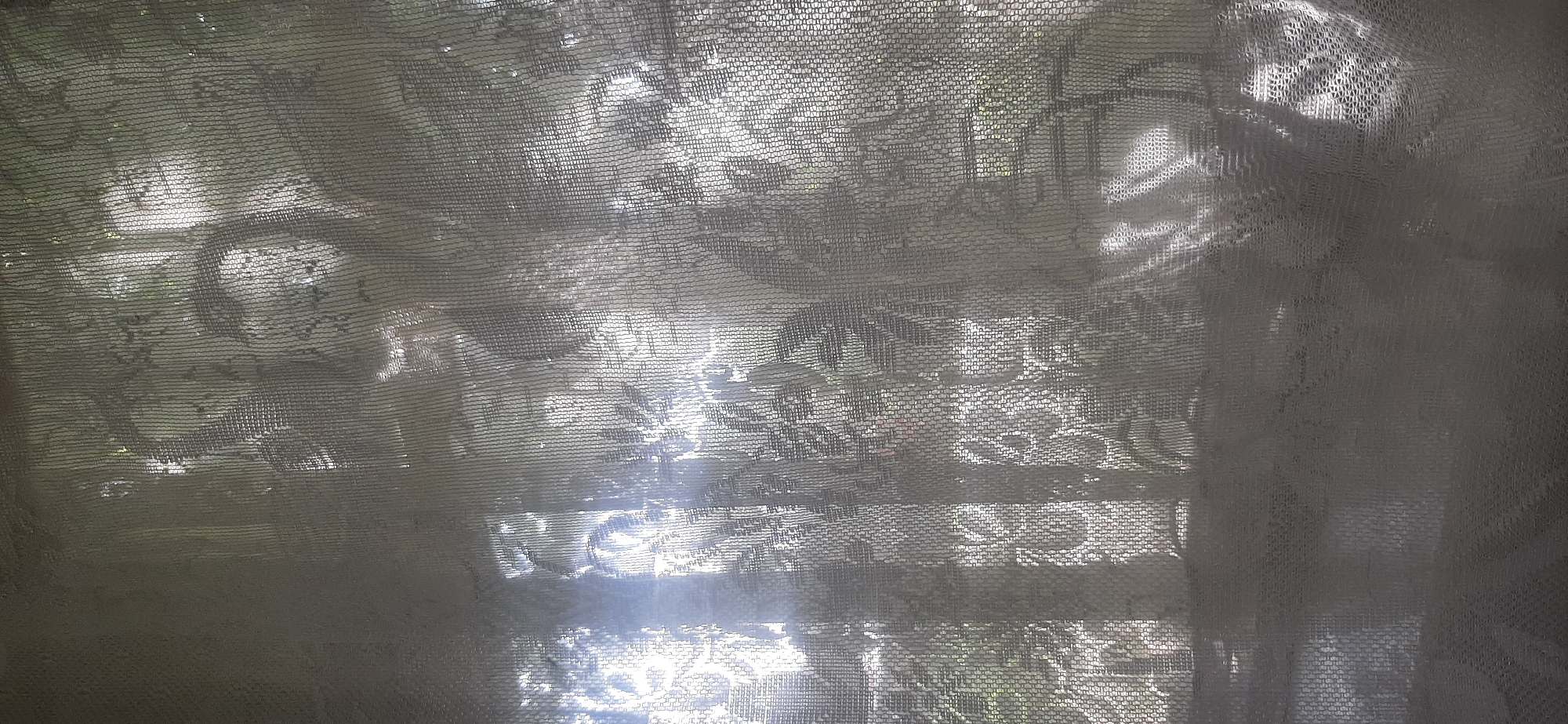 The image depicts a sheer, white lace curtain hanging over a window, through which sunlight filters. The sunlight creates dappled patterns and illuminates the intricate floral design of the lace. Beyond the curtain, the indistinct shapes of greenery, likely trees or bushes, are visible, suggesting an outdoor environment. The lighting is bright, with some areas appearing washed out due to the intensity of the sun. The overall impression is one of a domestic interior seen through a decorative textile. There is no information about the city of Yessentukskaya, Russia, within the image itself.